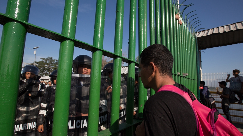 A Honduran migrant looks through the fence at military police in riot gear along the Guatemala-Mexico border