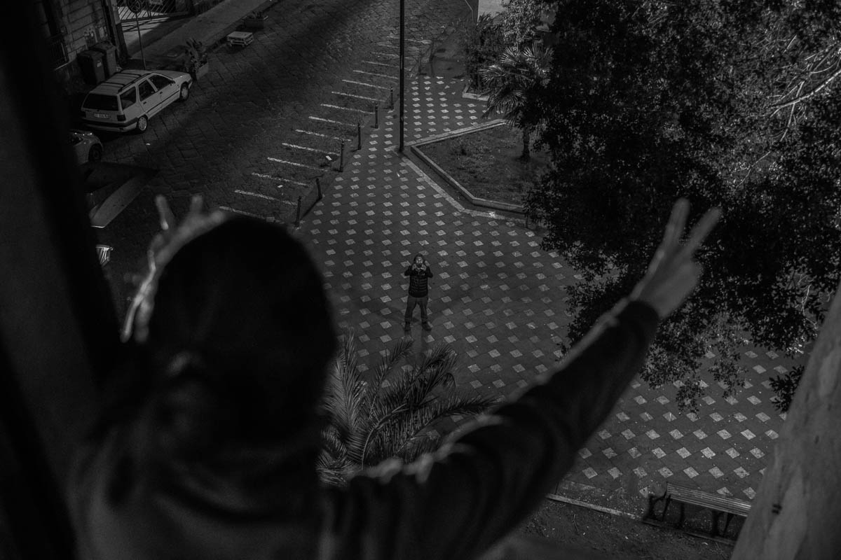 Marta greets from the window a friend who has just brought her home some food and left behind the door. It is 7 in the evening and there is no one else on the street. Marta greets him with a ''V'' victo