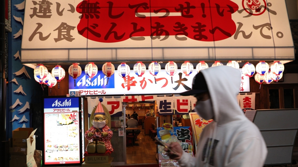 A man wearing a face mask walks past a restaurant illuminated at night in the Shinsekai shopping district of Osaka, Japan