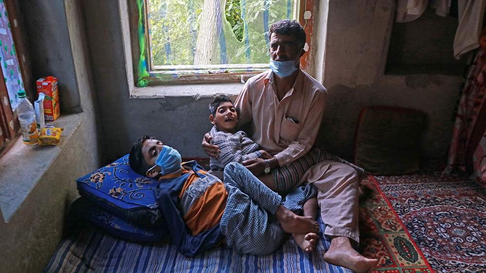 Nazir Ahmad Sheikh, a bus-conductor, at his home in Budgam village holding his paralysed son in his arms. Nazir has been left jobless due to the second consecutive lockdown in the region which marks a year on August 5