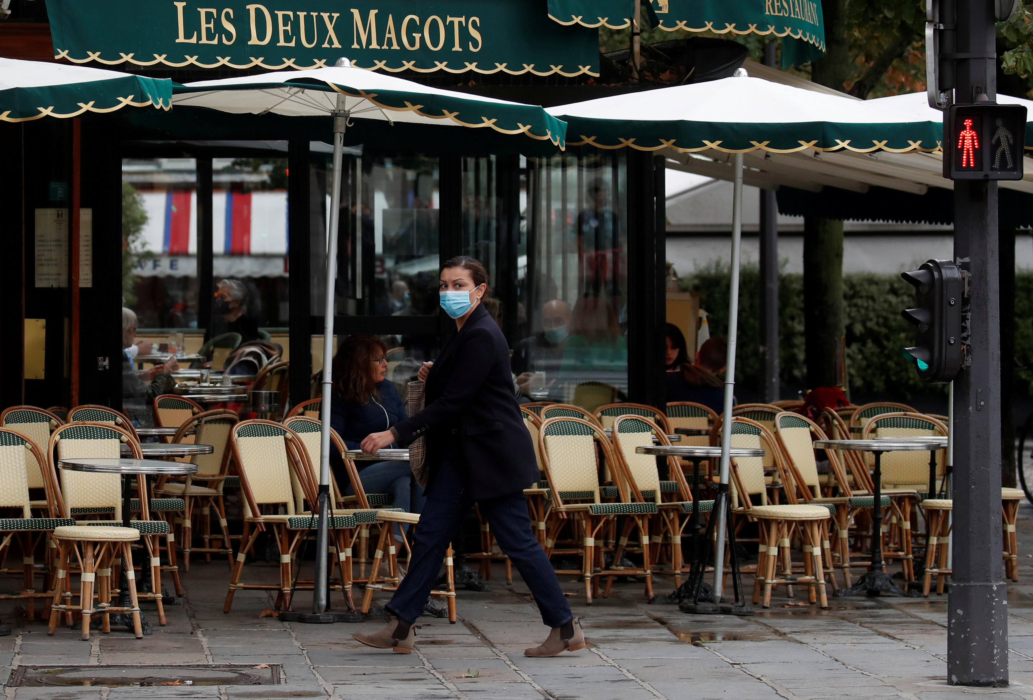 A woman wearing a protective face mask walks past a cafe in Paris [File: Gonzalo Fuentes/Reuters]