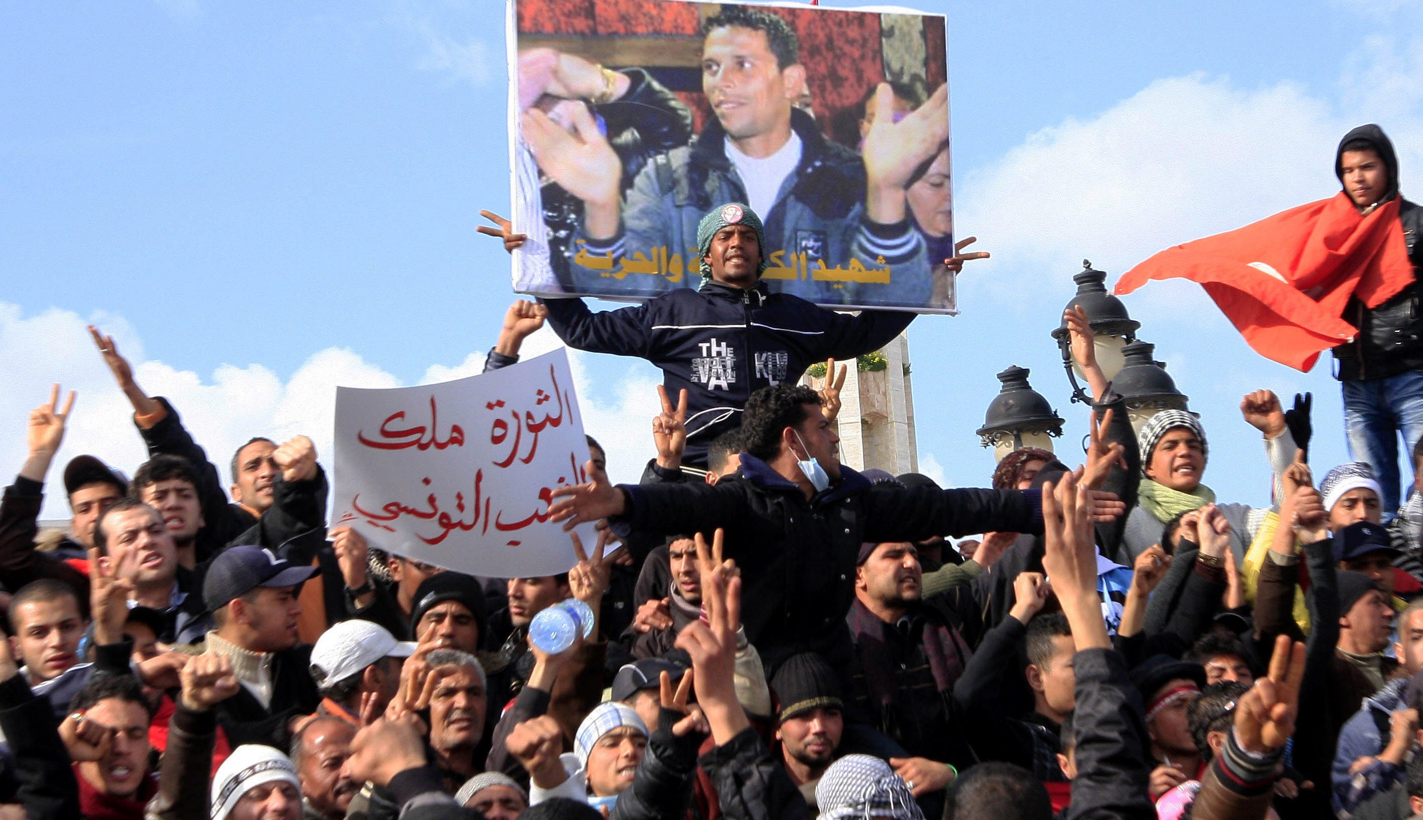 Tunisian protesters demonstrate beneath a poster of Mohamed Bouazizi, a street vendor who set himself on fire to protest against social conditions, starting the Arab Spring in late 2010 [File: Salah Habibi/AP]