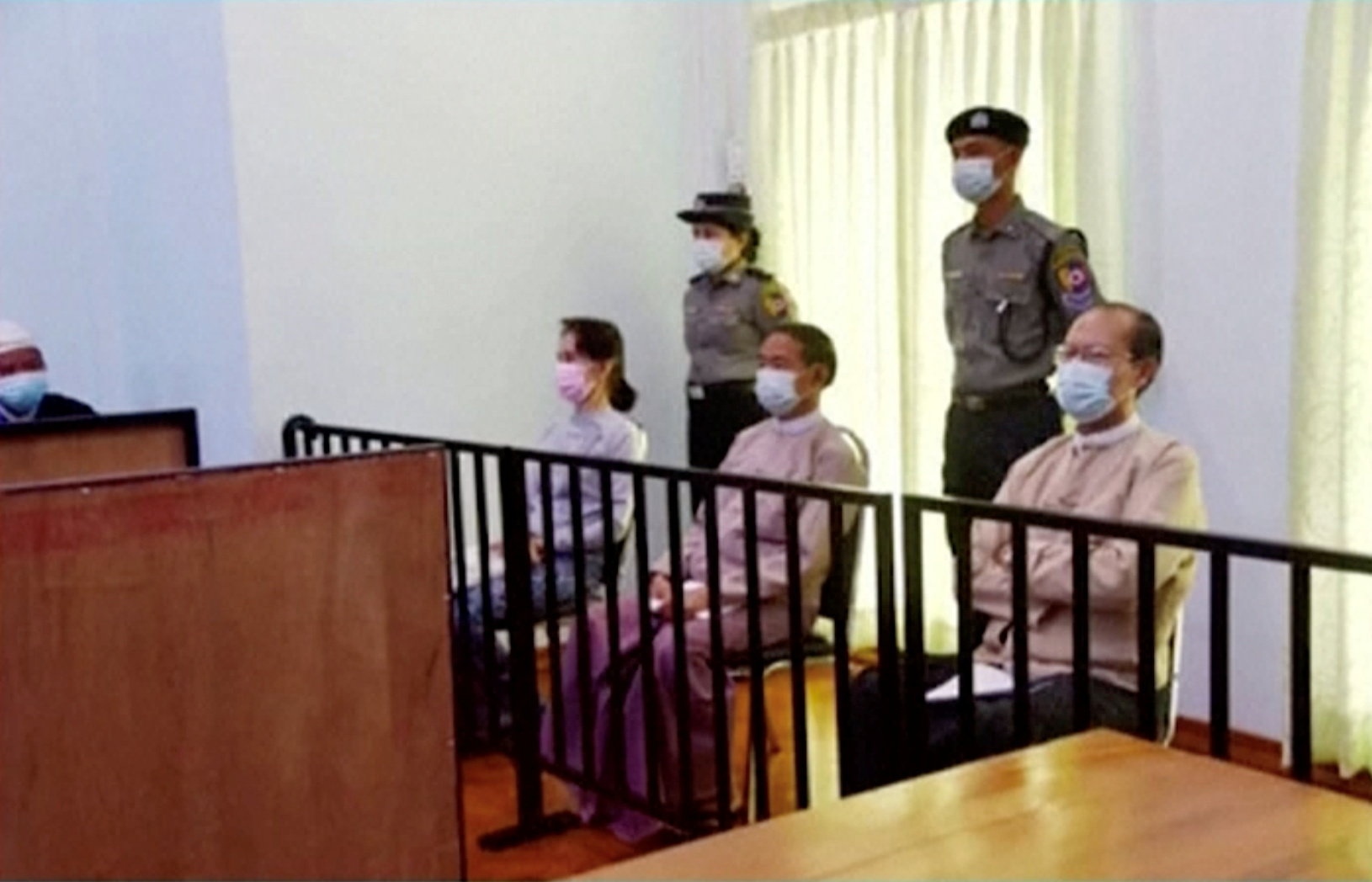 Aung San Suu Kyi sits in court with a female police officer behind her in her first public appearance since the February 1 coup