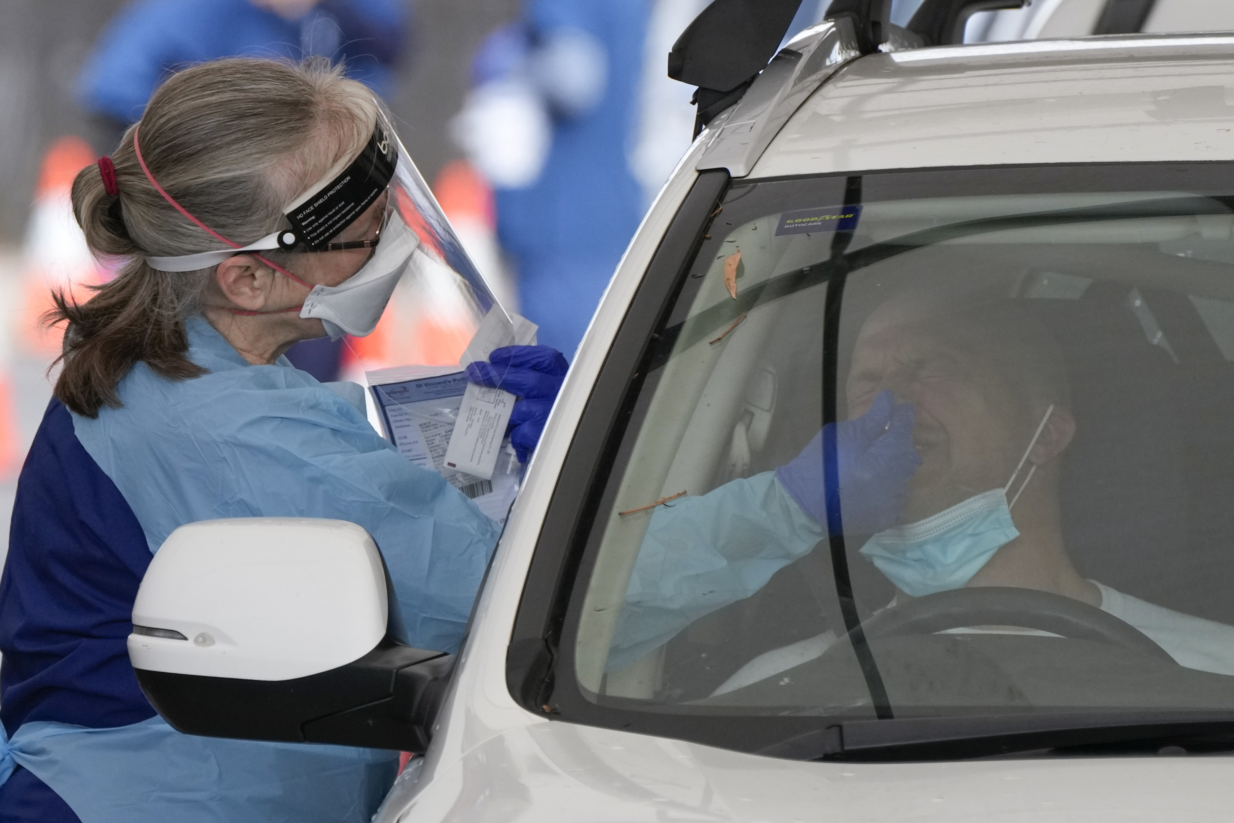 A woman in protective clothing administers a COVID test to someone sitting in a car