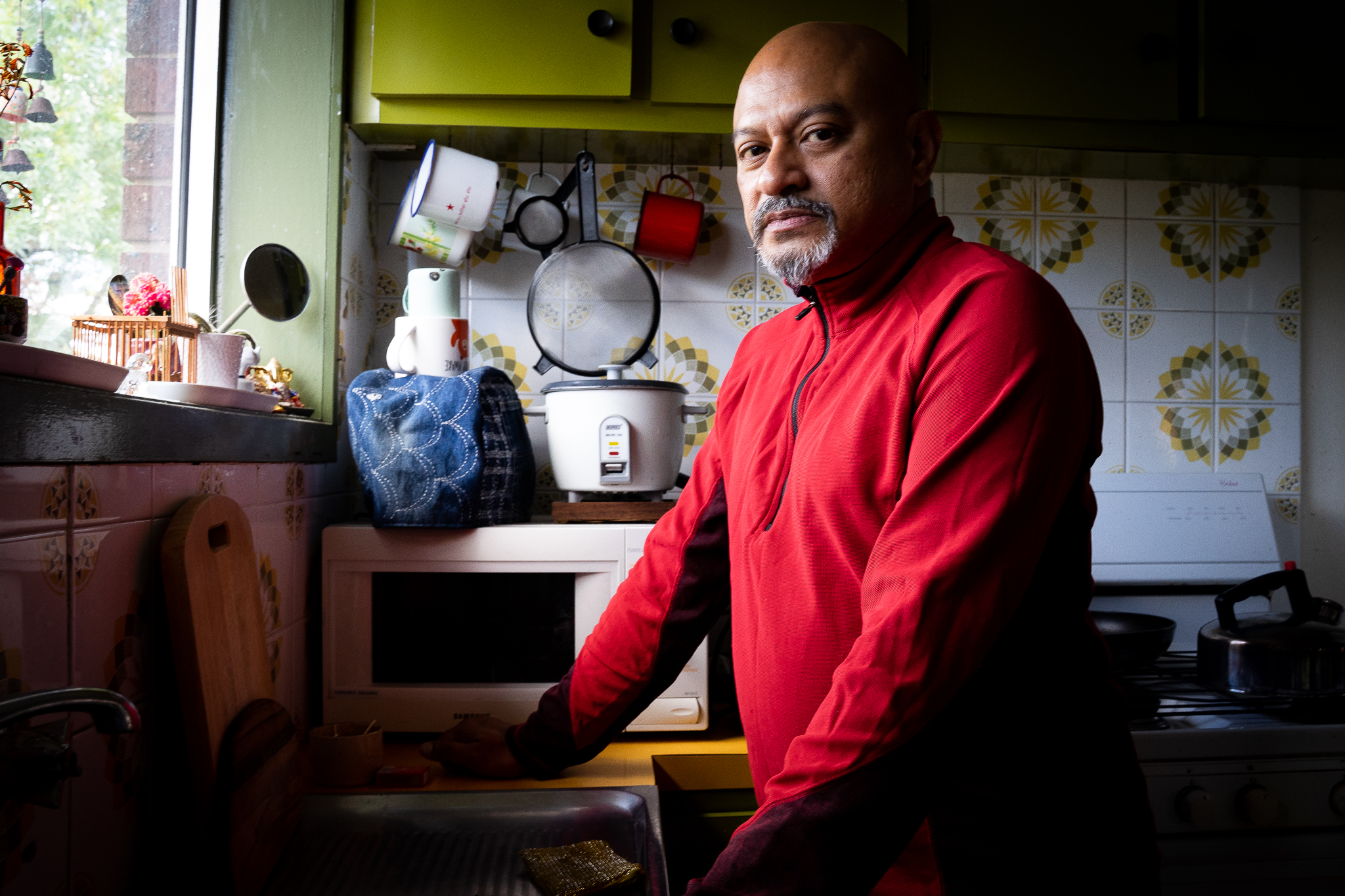 A portrait of Ian Row wearing a red sweater and standing in his kitchen
