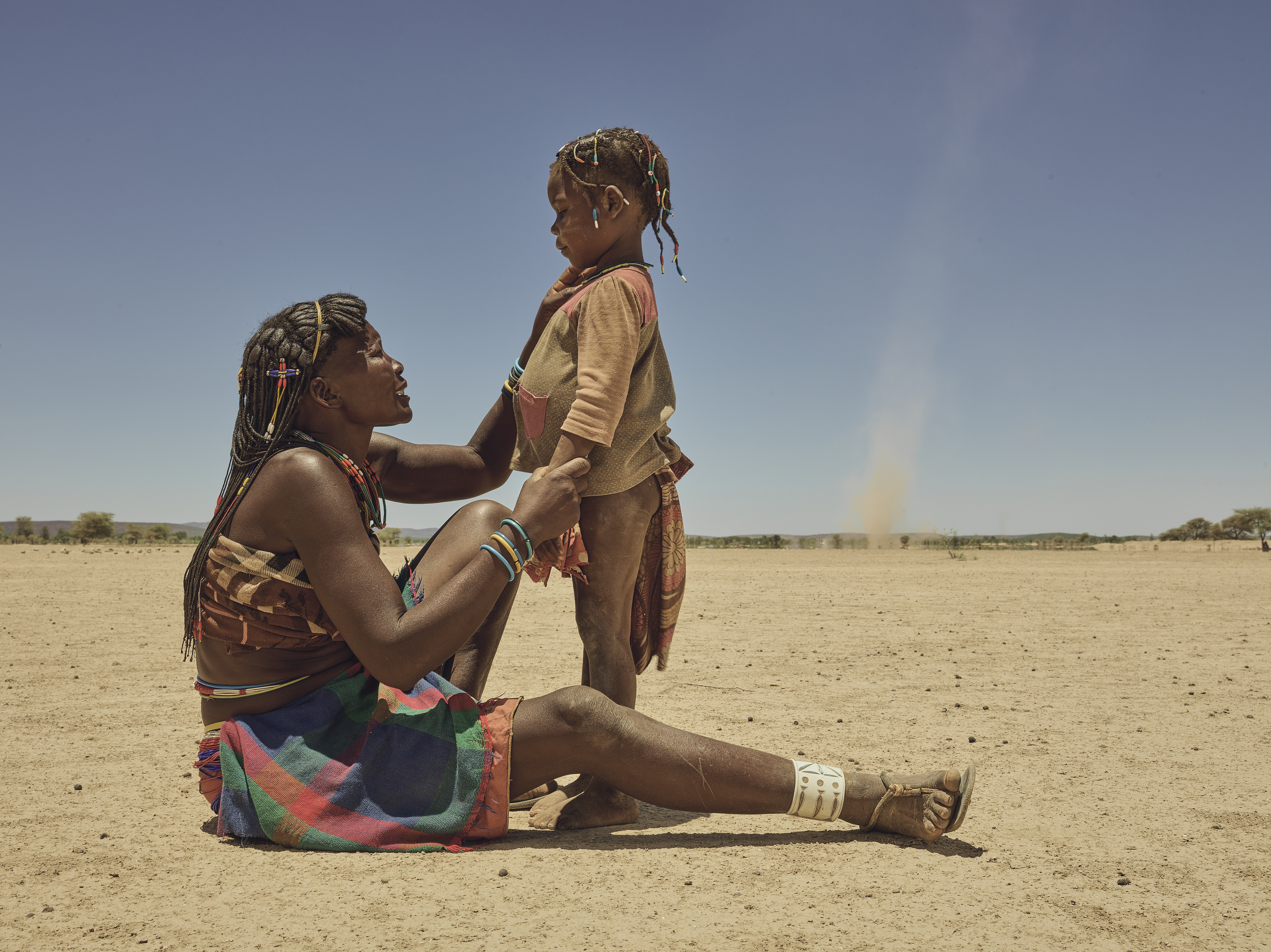 A photo of a woman sitting on the ground outside holding a standing child in front of her.