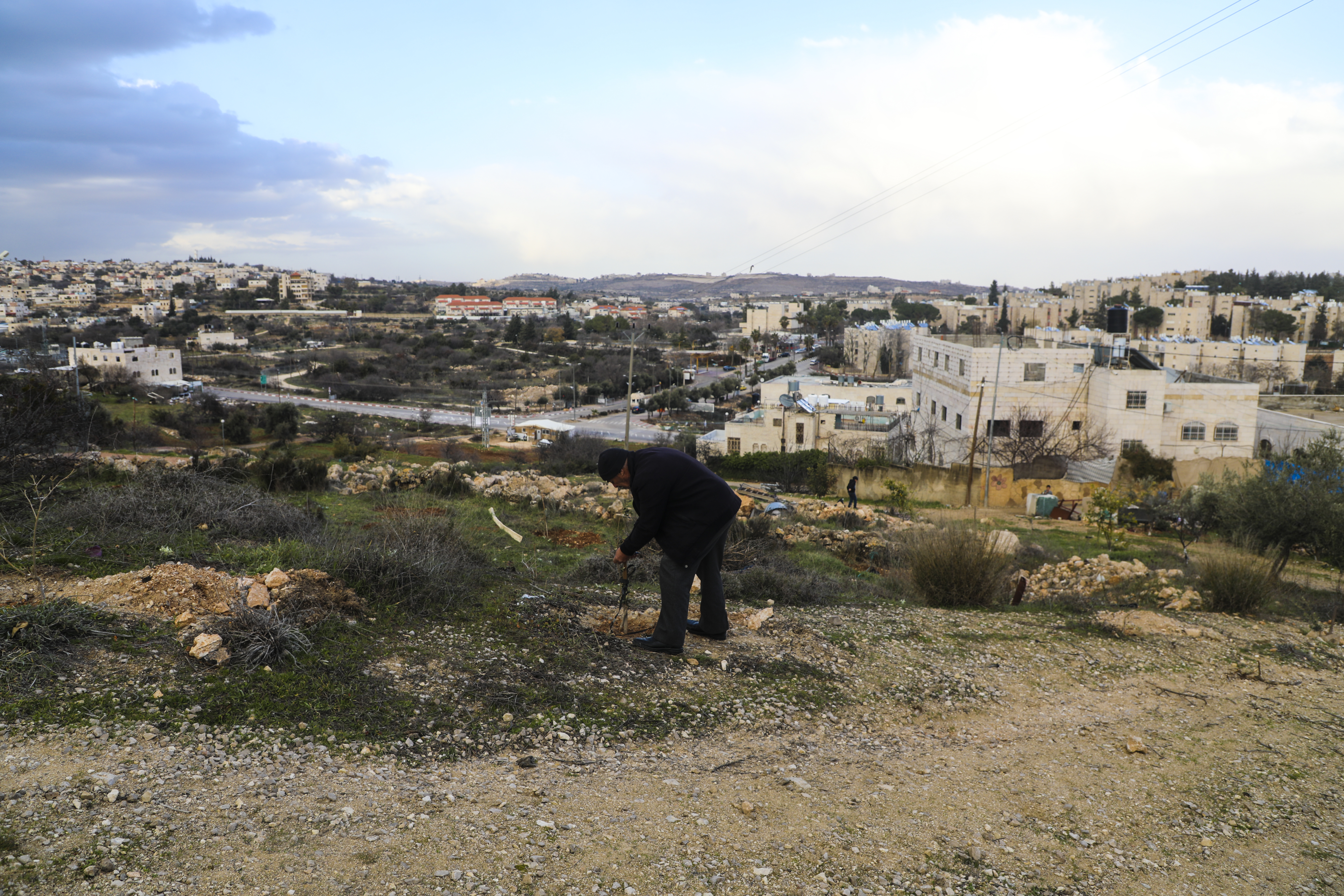 Abdulkareem al-Jaabari stands on his land with his home to the right, and the Israeli settlement of Kiryat Arbaa in the background