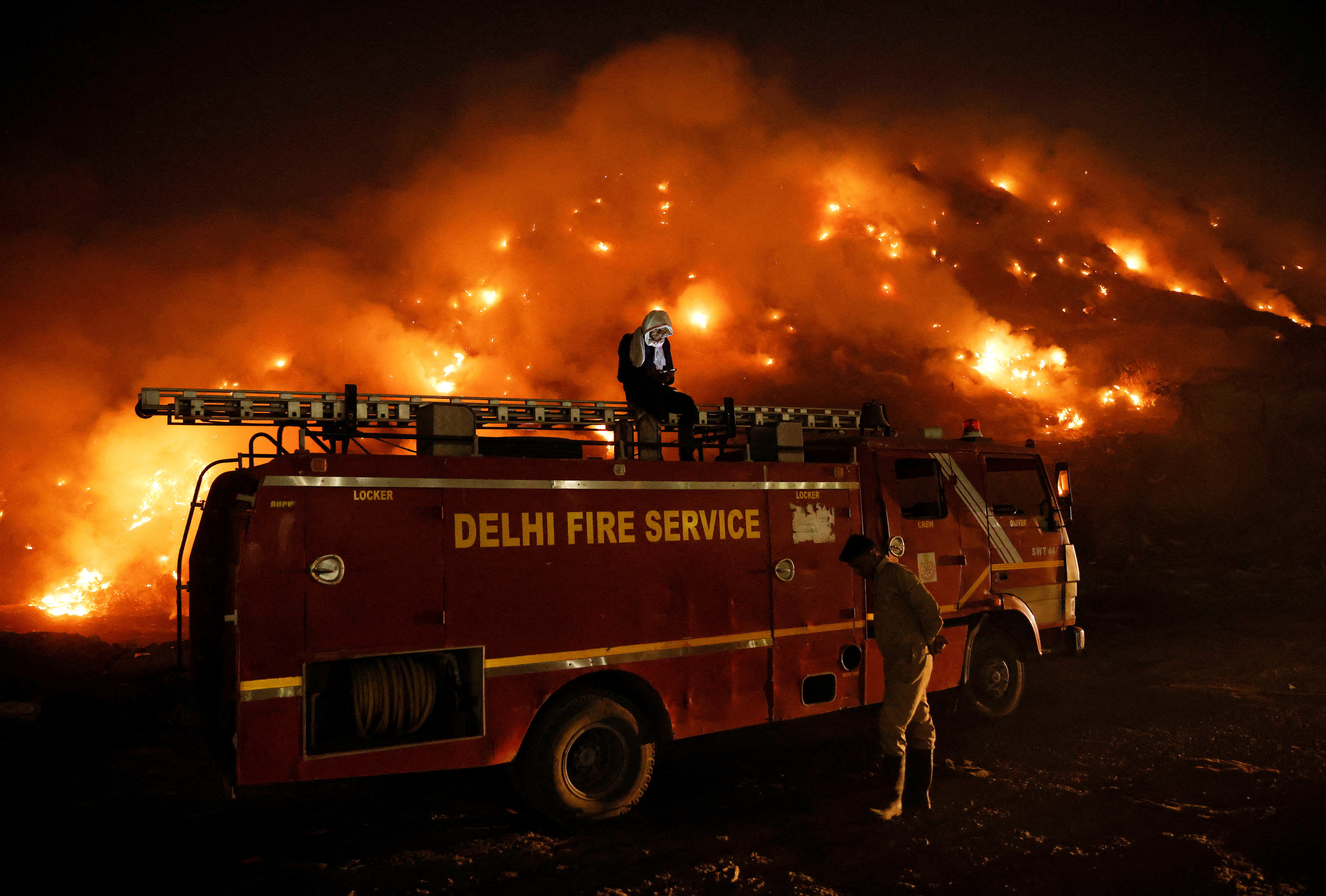 A firefighter uses his mobile phone as he sits on a fire truck as smoke billows from burning garbage at the Bhalswa landfill site in New Delhi