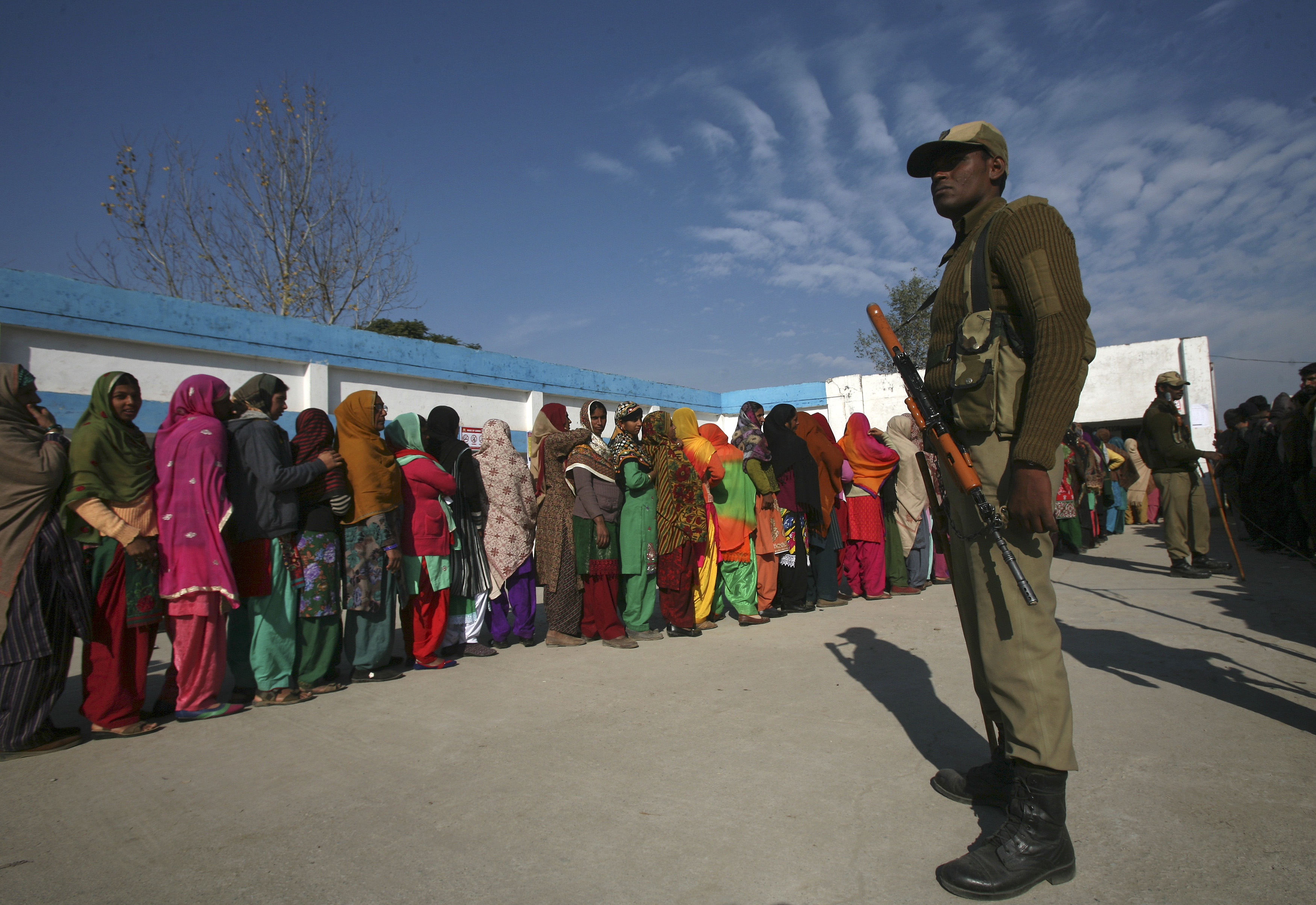 Voters line up to cast their votes as an Indian officer stands guard outside a polling station during assembly polls in Indian-administered Kashmir in 2014