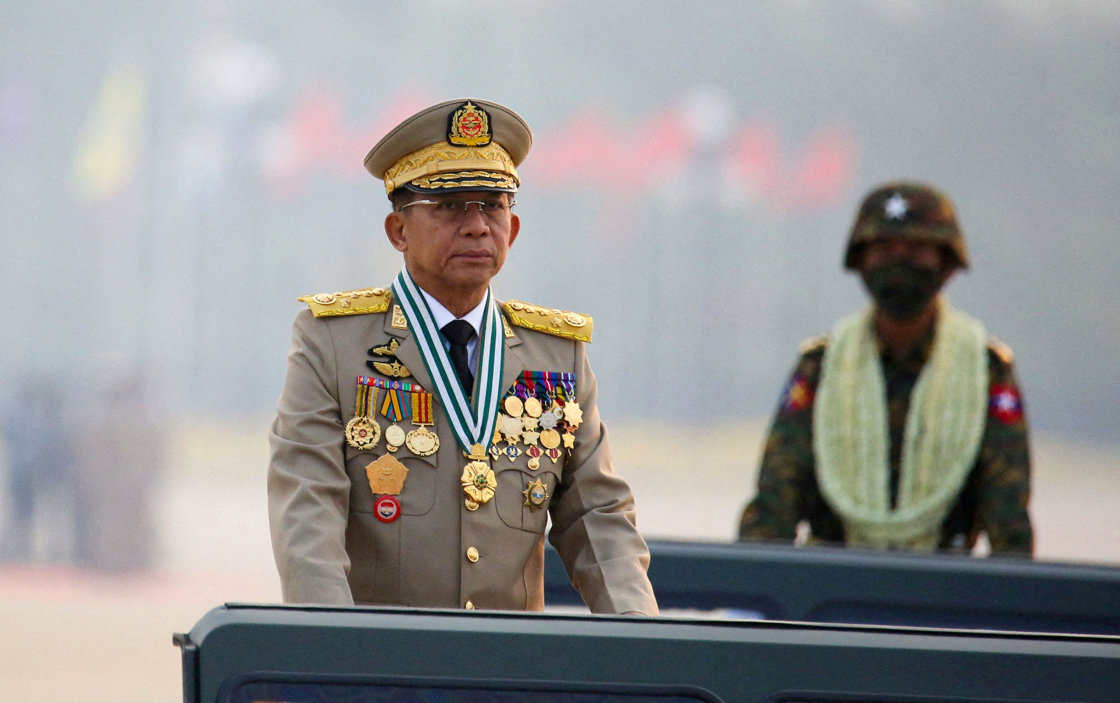 Min Aung Hlaing in full military regalia rides in the back of a jeep to inspect the forces with a soldier in fatigues in the vehicle behind.