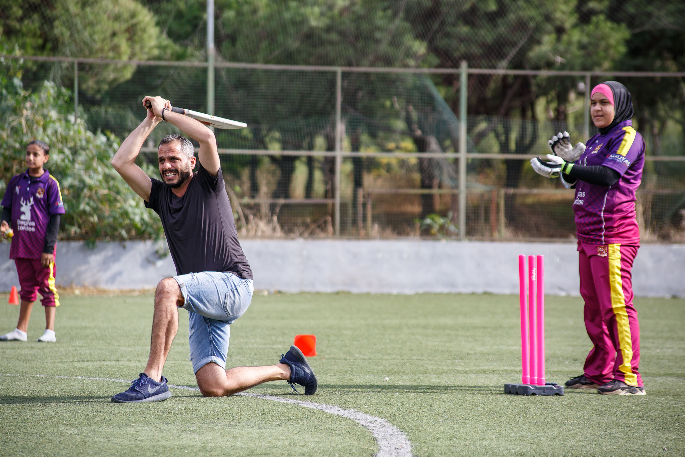 A coach swings a cricket bat on a field. A girl stands behind the wicket.