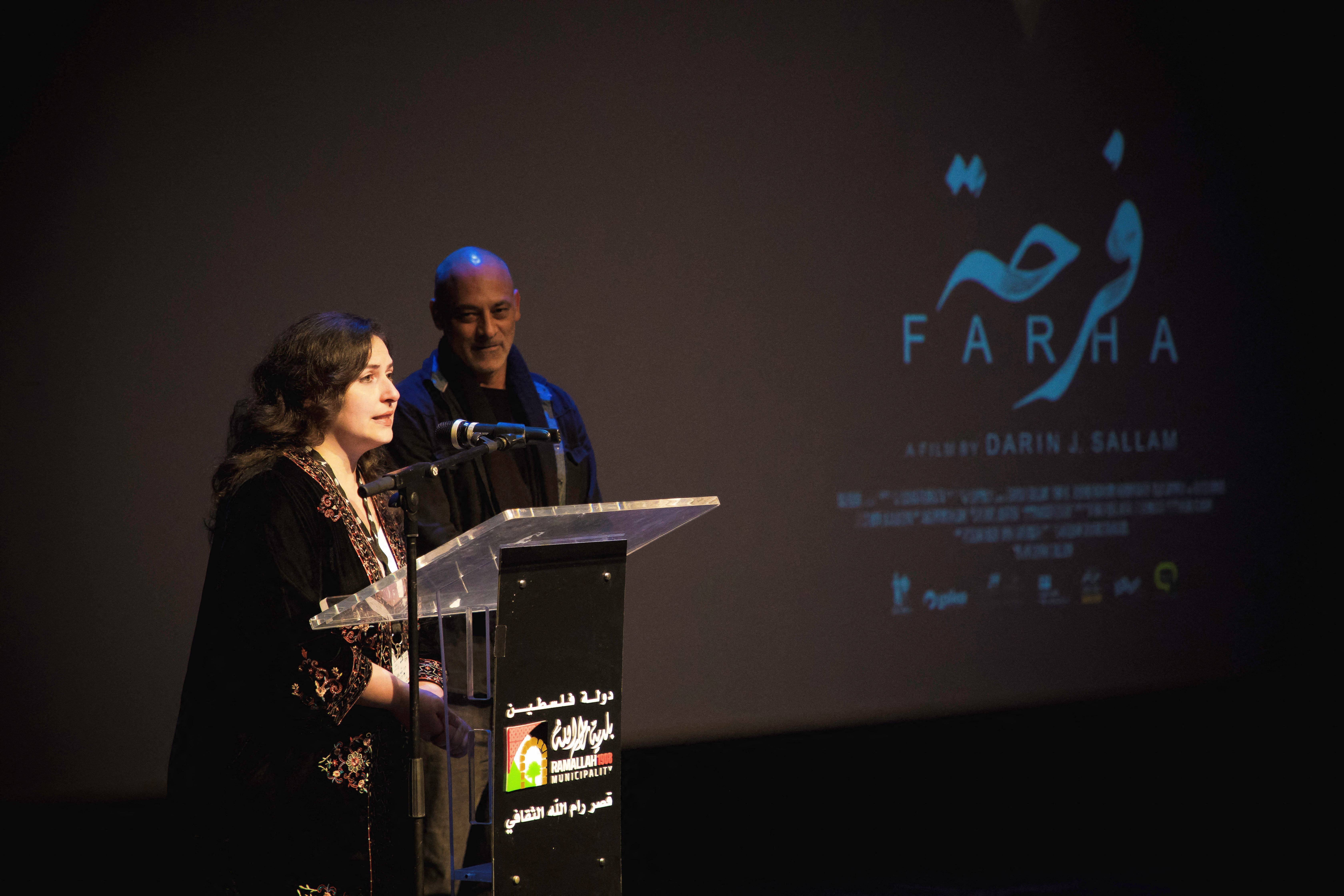 Woman and man stand next to a lecturn with 'Farha' written in Arabic on the backdrop