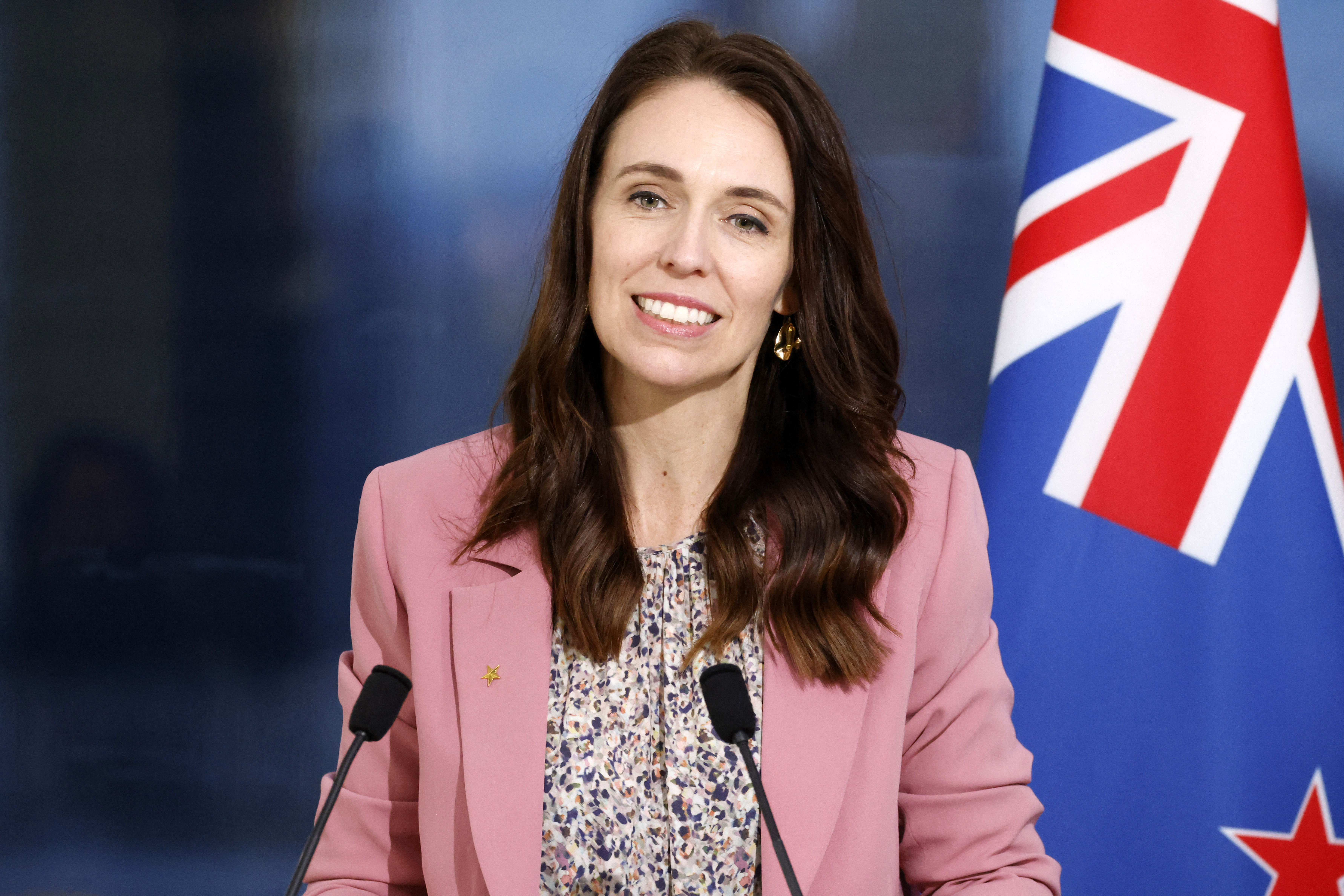 Ardern in a pink jacket smiles from behind two microphones with a New Zealand flag behind her