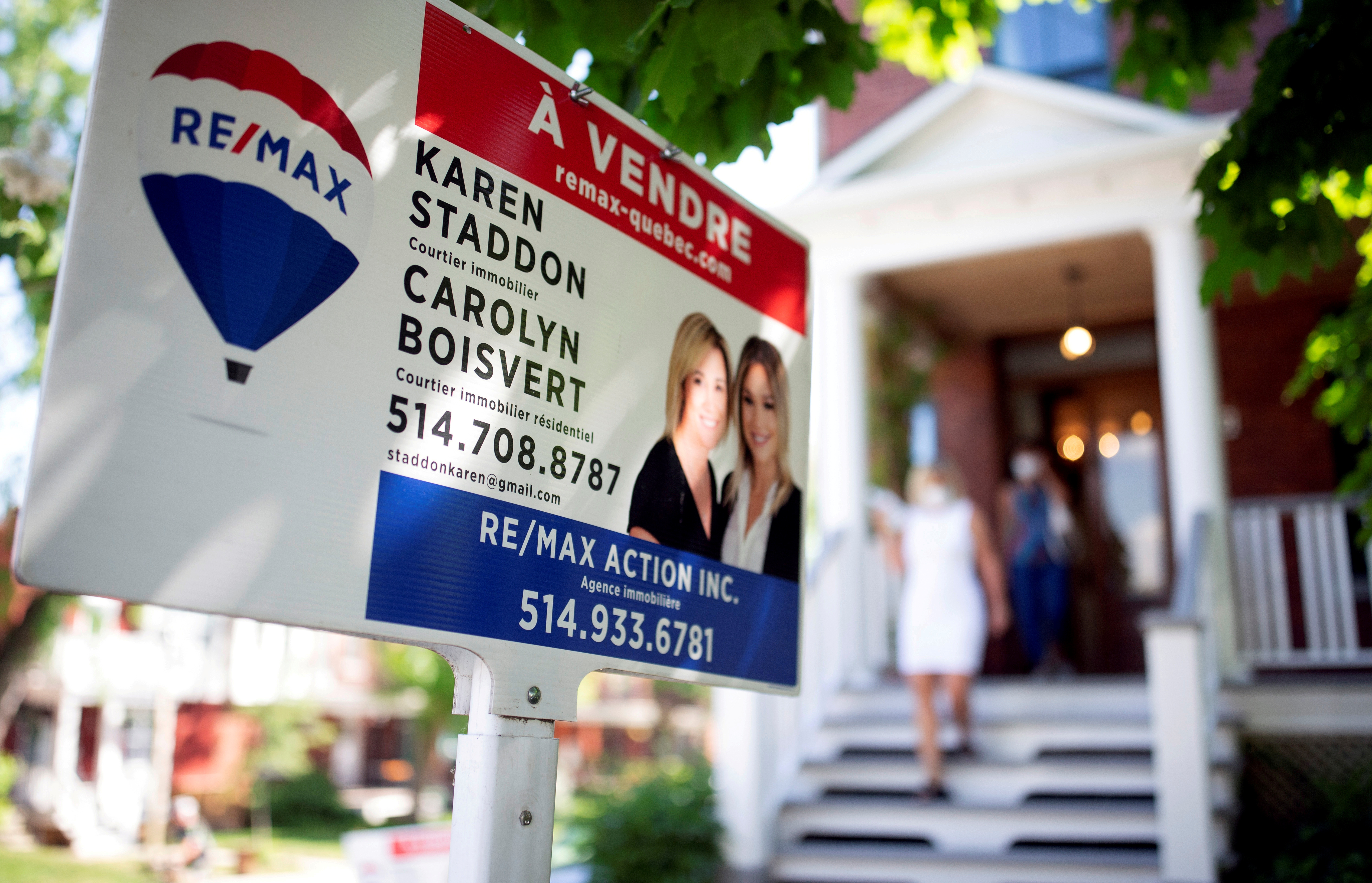 Close up of a For Sale sign outside. In the background is the blurred white house, with a woman walking up the wooden steps