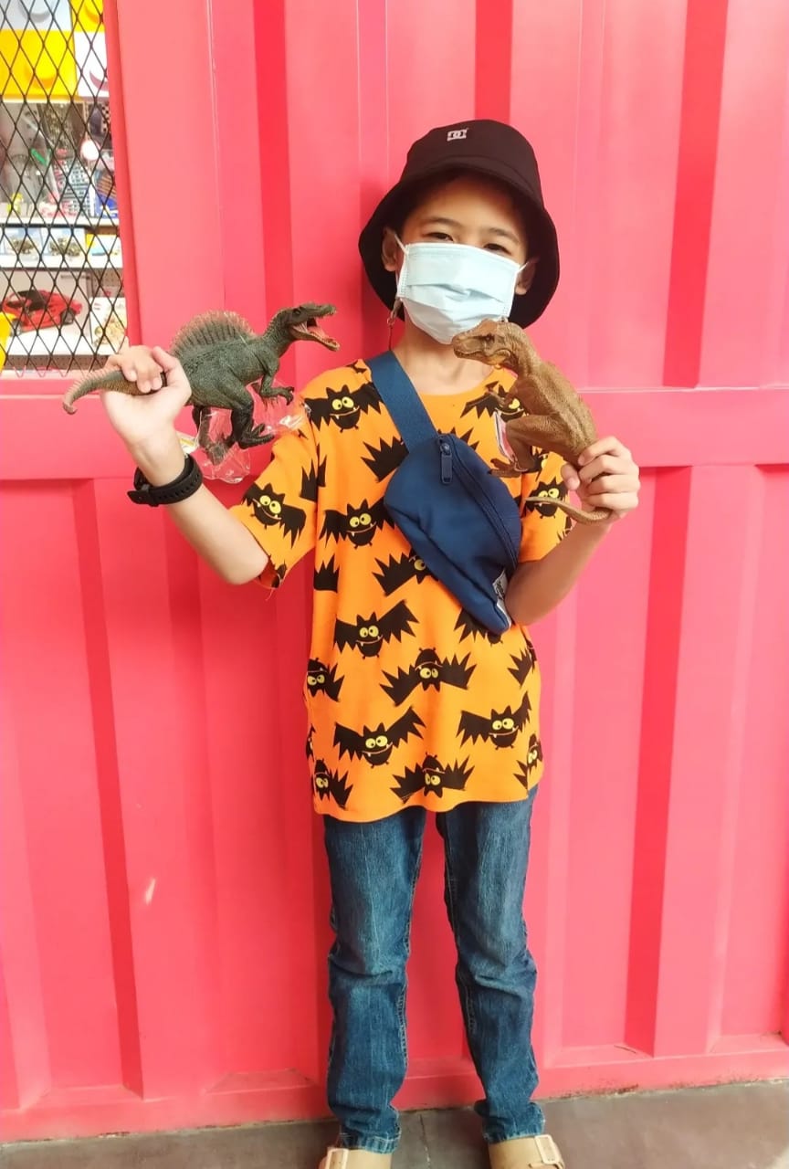 Panghegar standing in front of a red wall. He's wearing an orange t-shirt with a print of black cartoon bats and jeans. He's got a hat on his head and is holding a dinosaur toy in each of his hands.