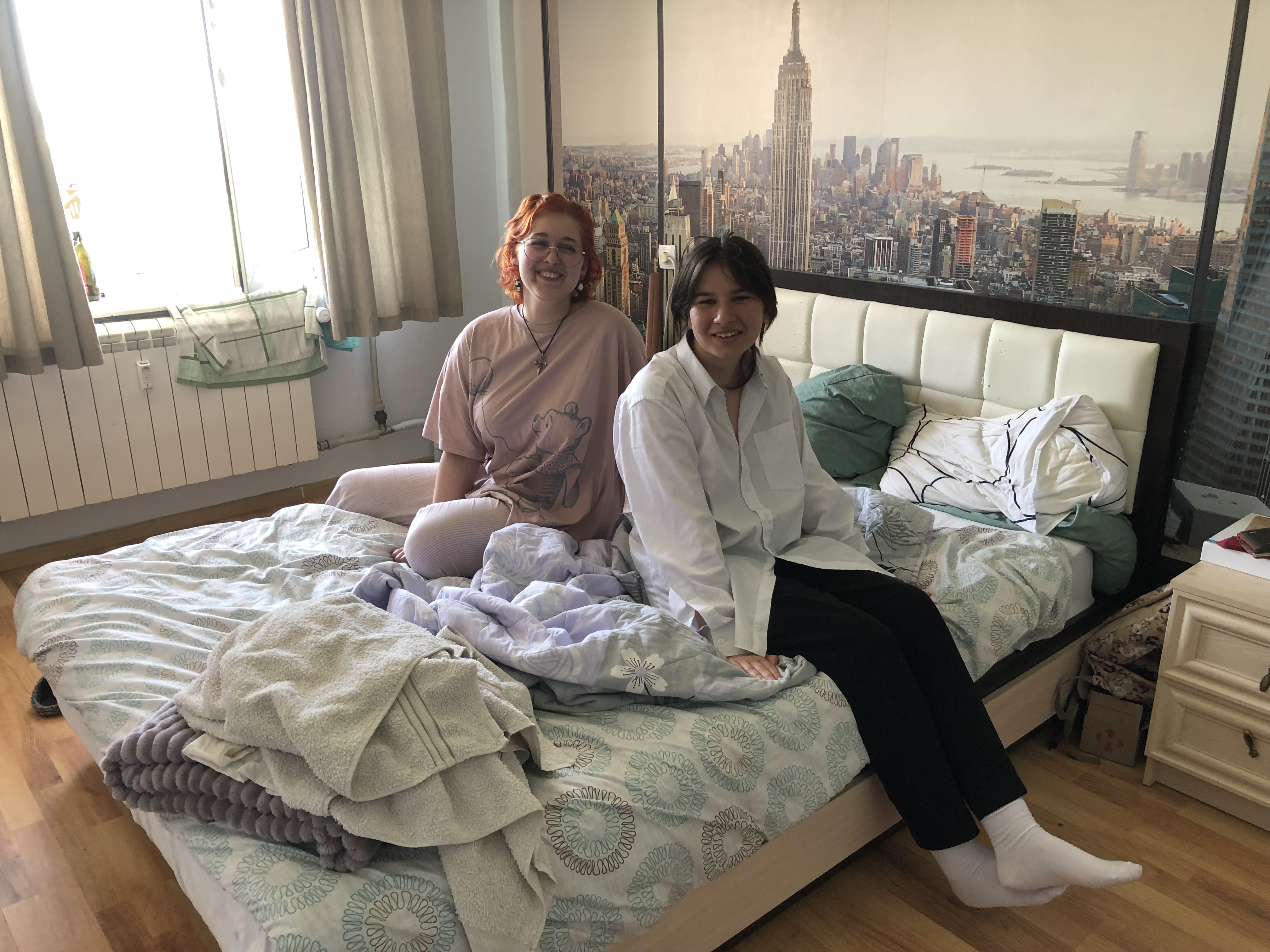 Tanya and Veronika sitting on the bed they share in the apartment