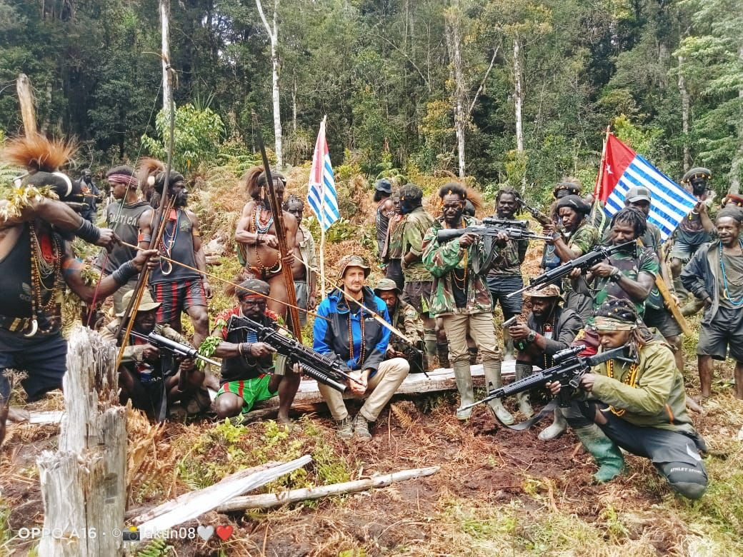 Philip Mehrtens sitting on a log in the forest with the group of TPN-PB fighters who took him hostage in February, Thet have weapons and there are Papuan flags. The pictures was released in March.