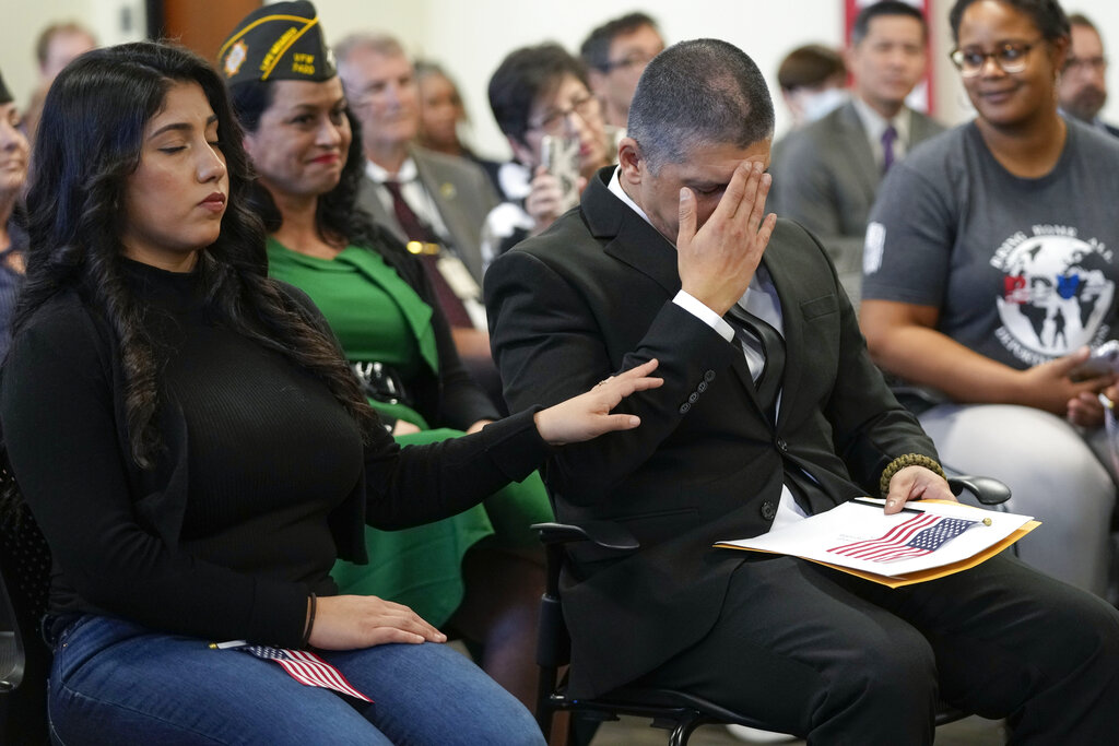 Mata in a black suit, sitting near his wife, has his head in his hand looking emotional as his wife rests her hand on his shoulder in comfort