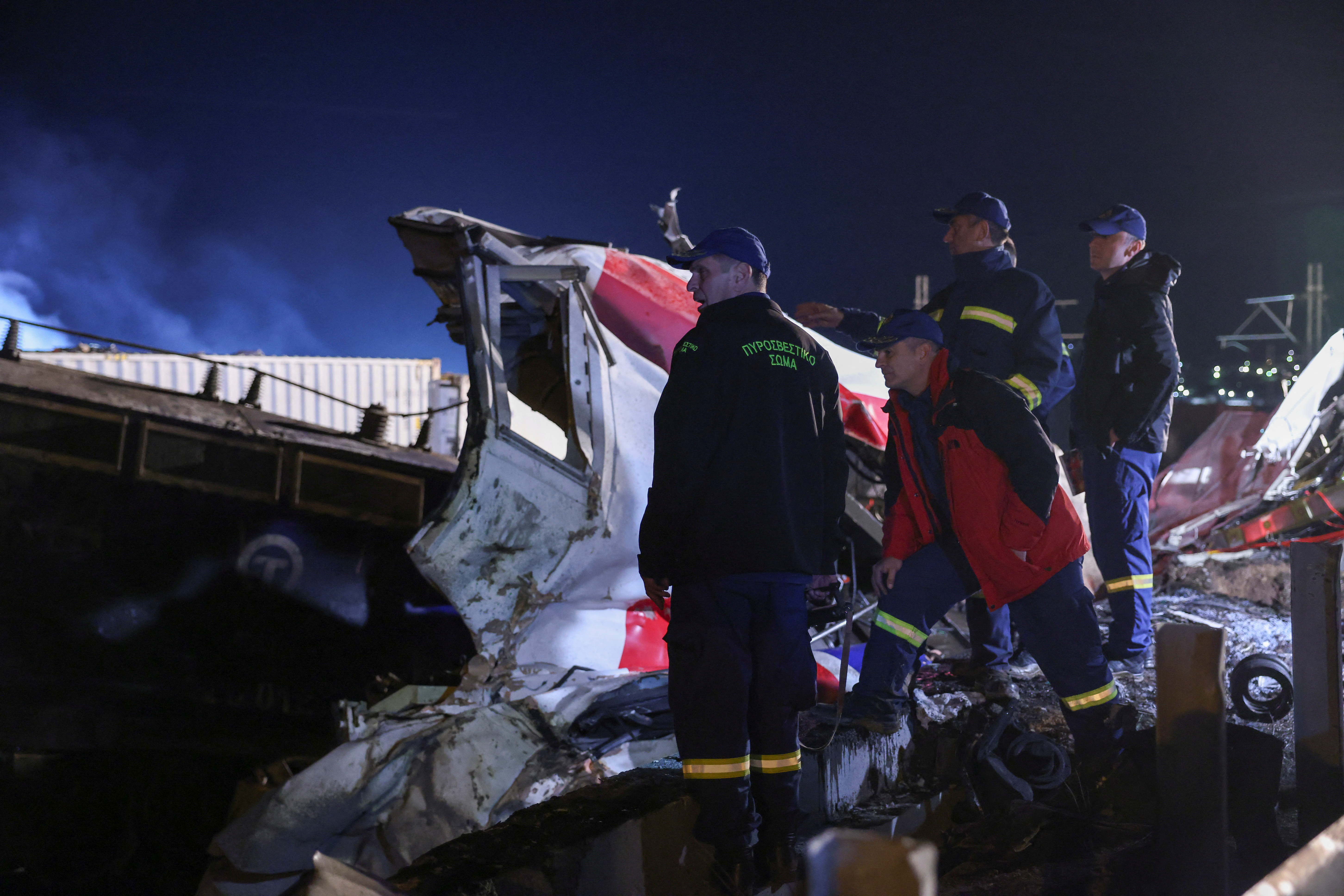 Firefighters at the site of crash near Larissa. There is a mangled red and white train car behind them. 