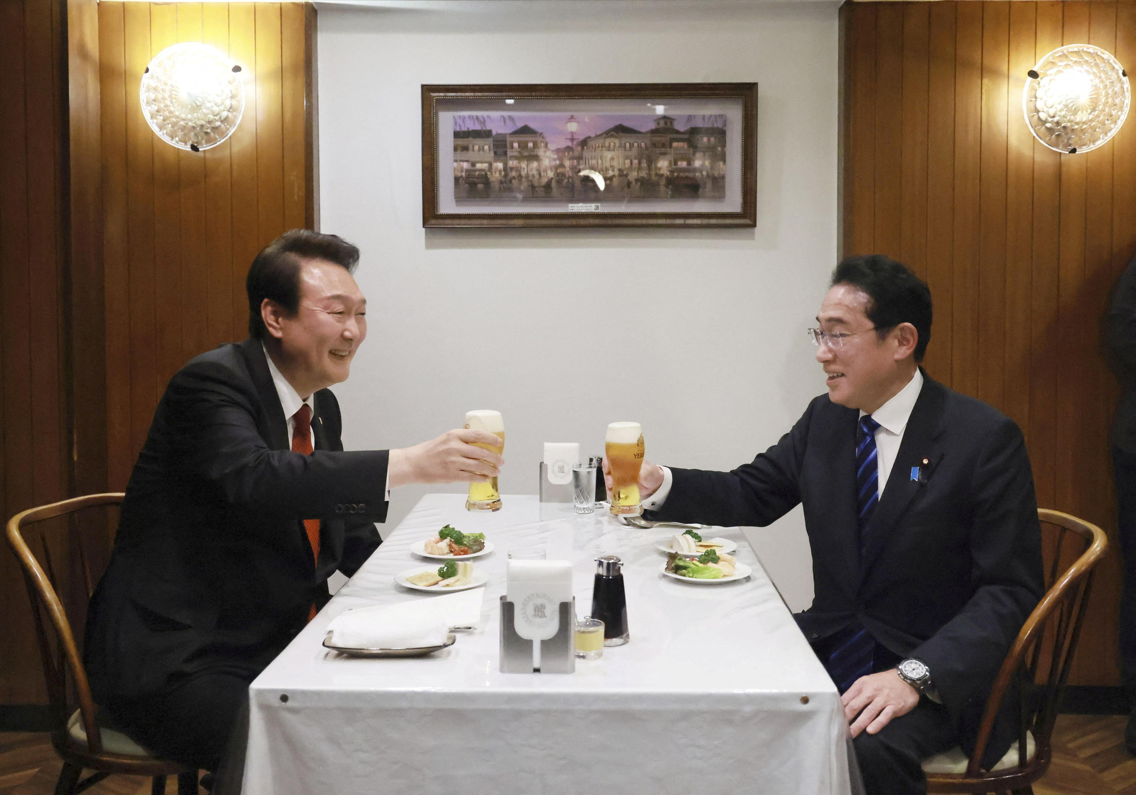 Fumio Kishida (right) and Yoon Suk-yeol toast with a glass of beer during a meal at a Tokyo restaurant. They are sitting opposite each other at a table. There are some appetisers in front of them. They are smiling.
