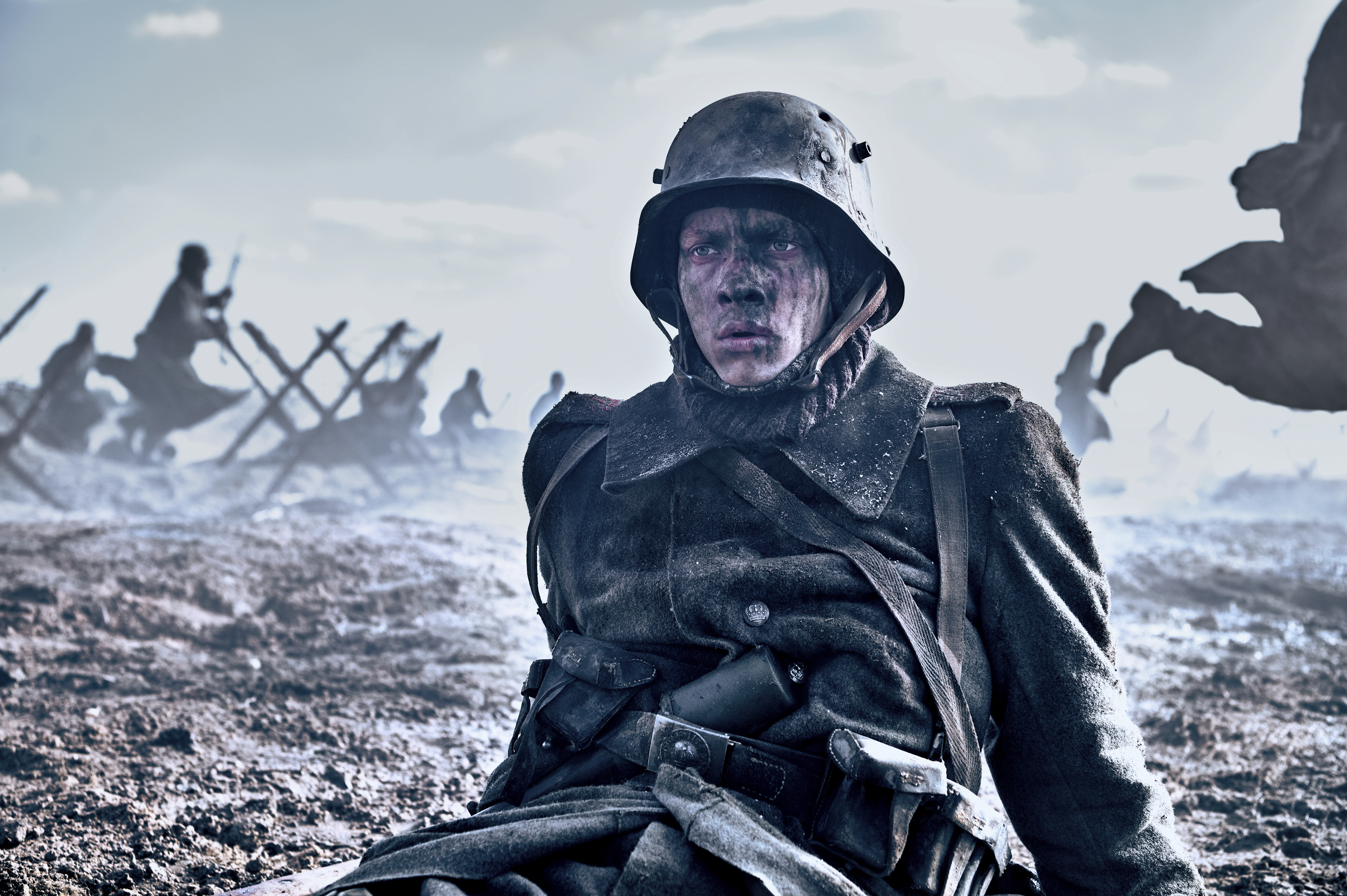 A man in a World War I uniform, covered in mud, lies on the ground as soldiers run behind him