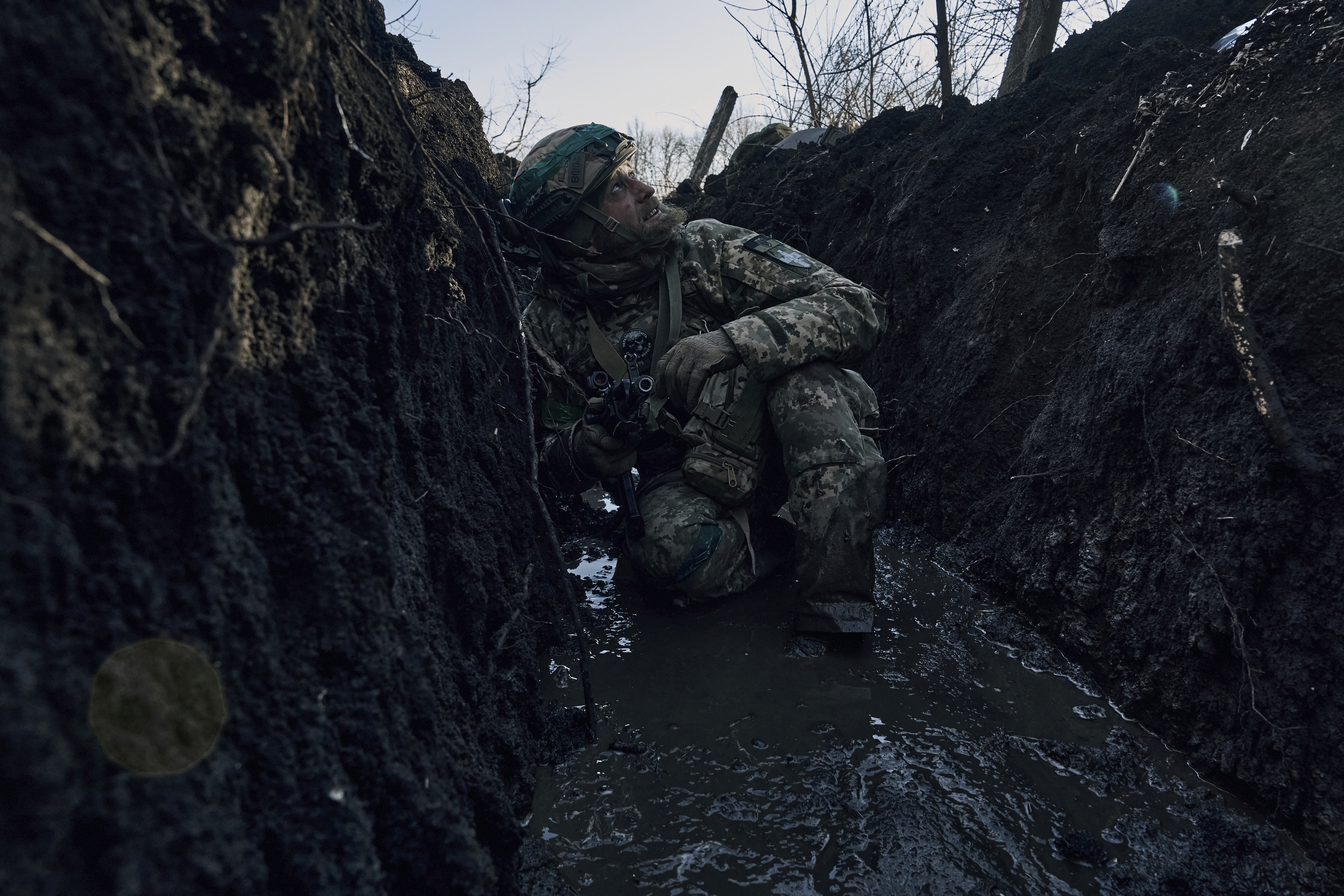 A Ukrainian soldier crouches down in a trench near the front line in Bakhmut, Ukraine. He's looking to the sky and has his weapon in his right hand. It's very muddy.