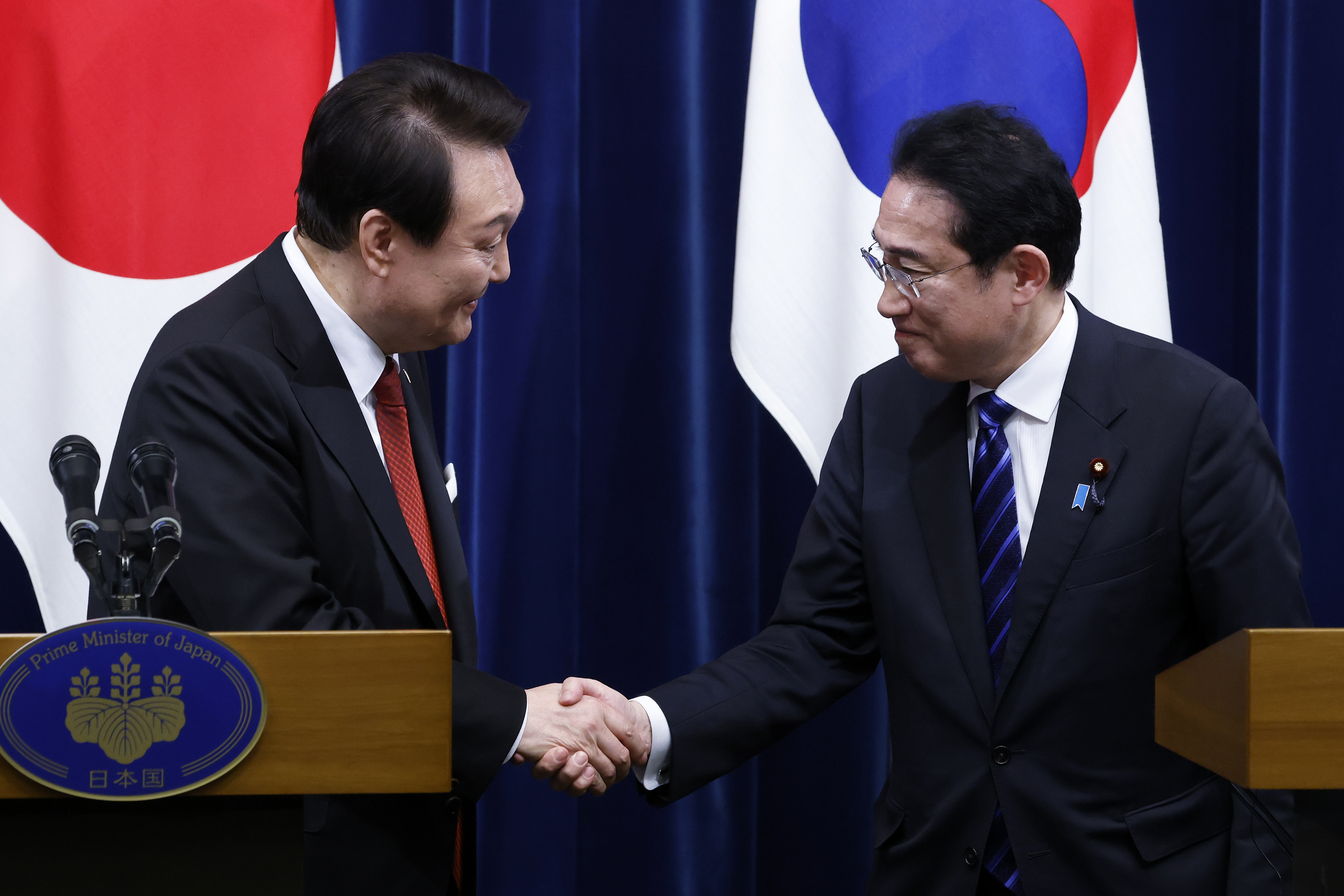 South Korea's Yoon Suk-yeok and Japan's Fumio Kishida shaking hands and smiling in front of the two countries' flags.