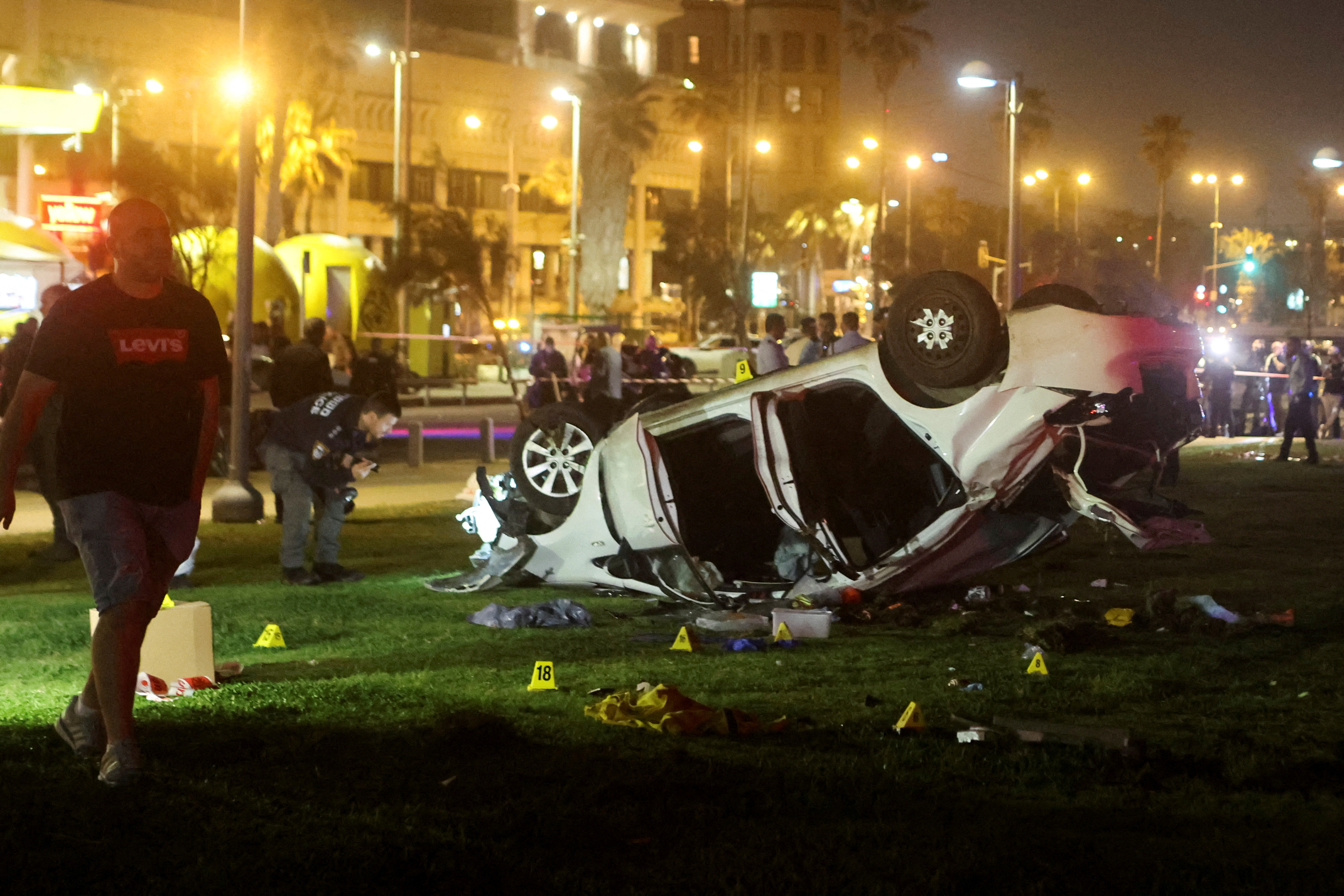 The scene of the attack in Tel Aviv. It is night time, there are lots of lights and a white car turned over with its doors open. There are people around.