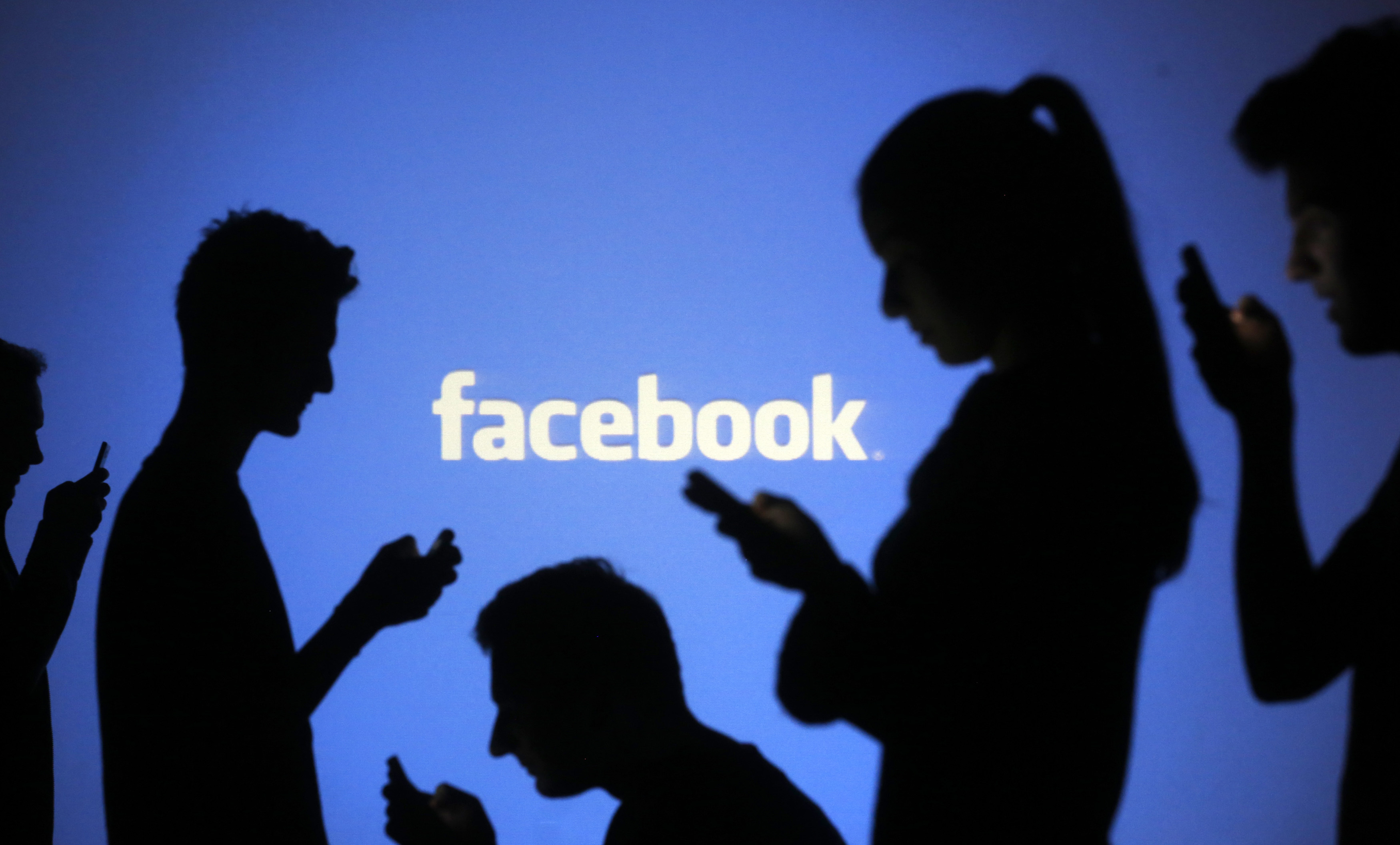People are silhouetted as they pose with mobile devices in front of a screen projected with a Facebook logo, in this picture illustration taken in Zenica October 29, 2014