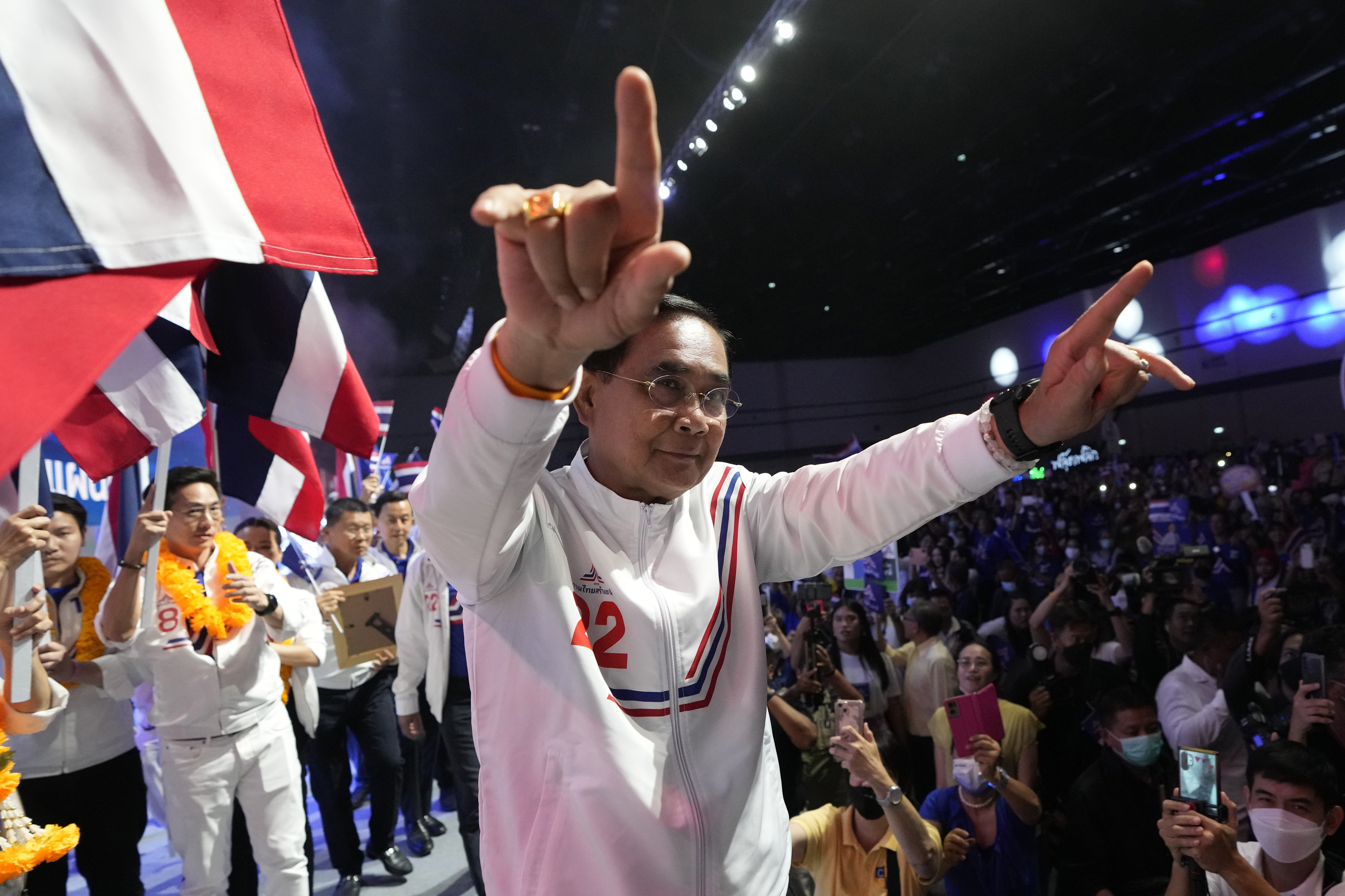 Prayuth gesturing to his supporters. He is wearing a track suit in the party colours of red white and blue and there are flags in the background