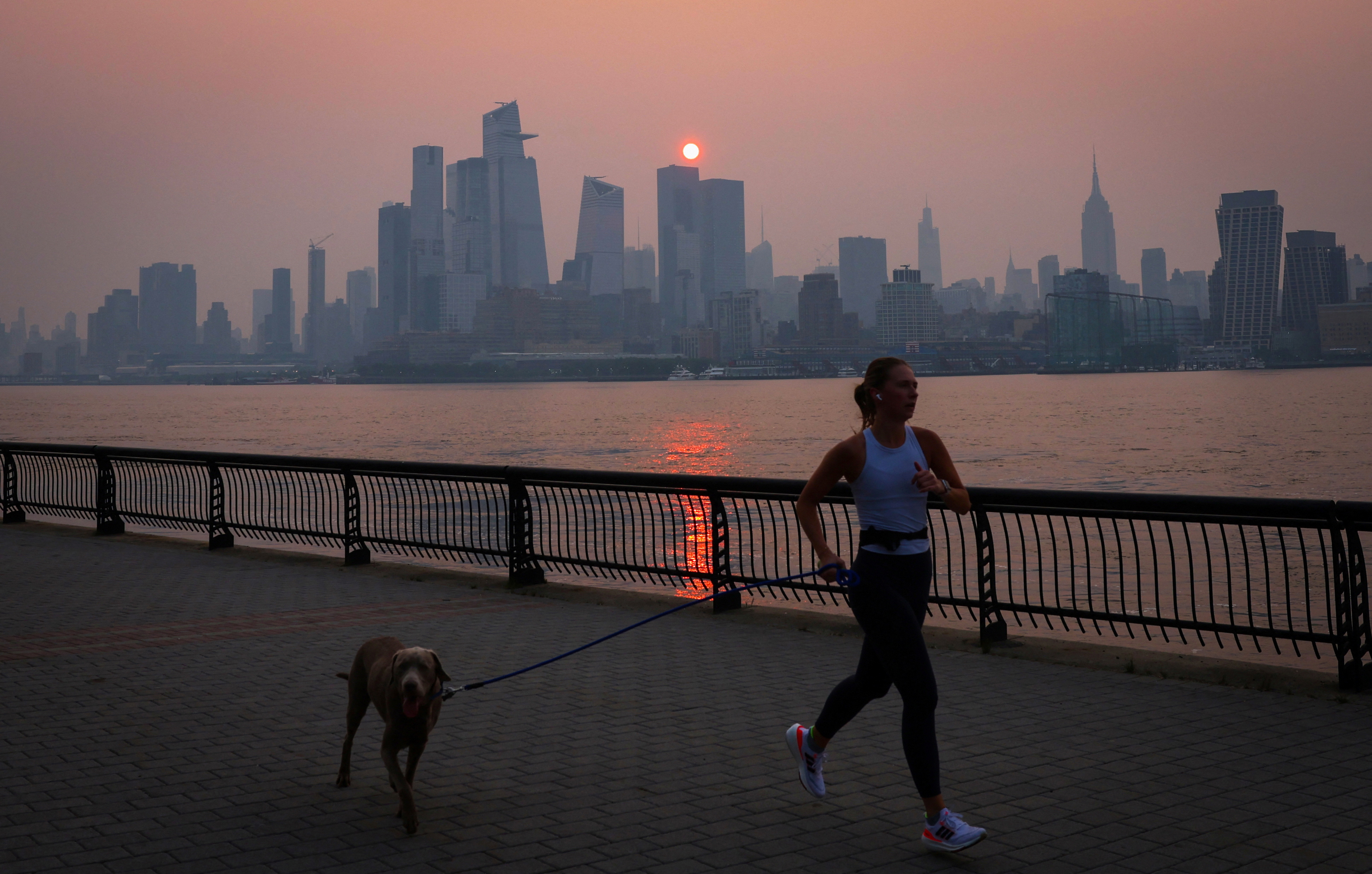A woman jogs along the Hudson River shortly after sunrise on June 7, 2023, as haze and smoke caused by wildfires in Canada hang over the Manhattan skyline