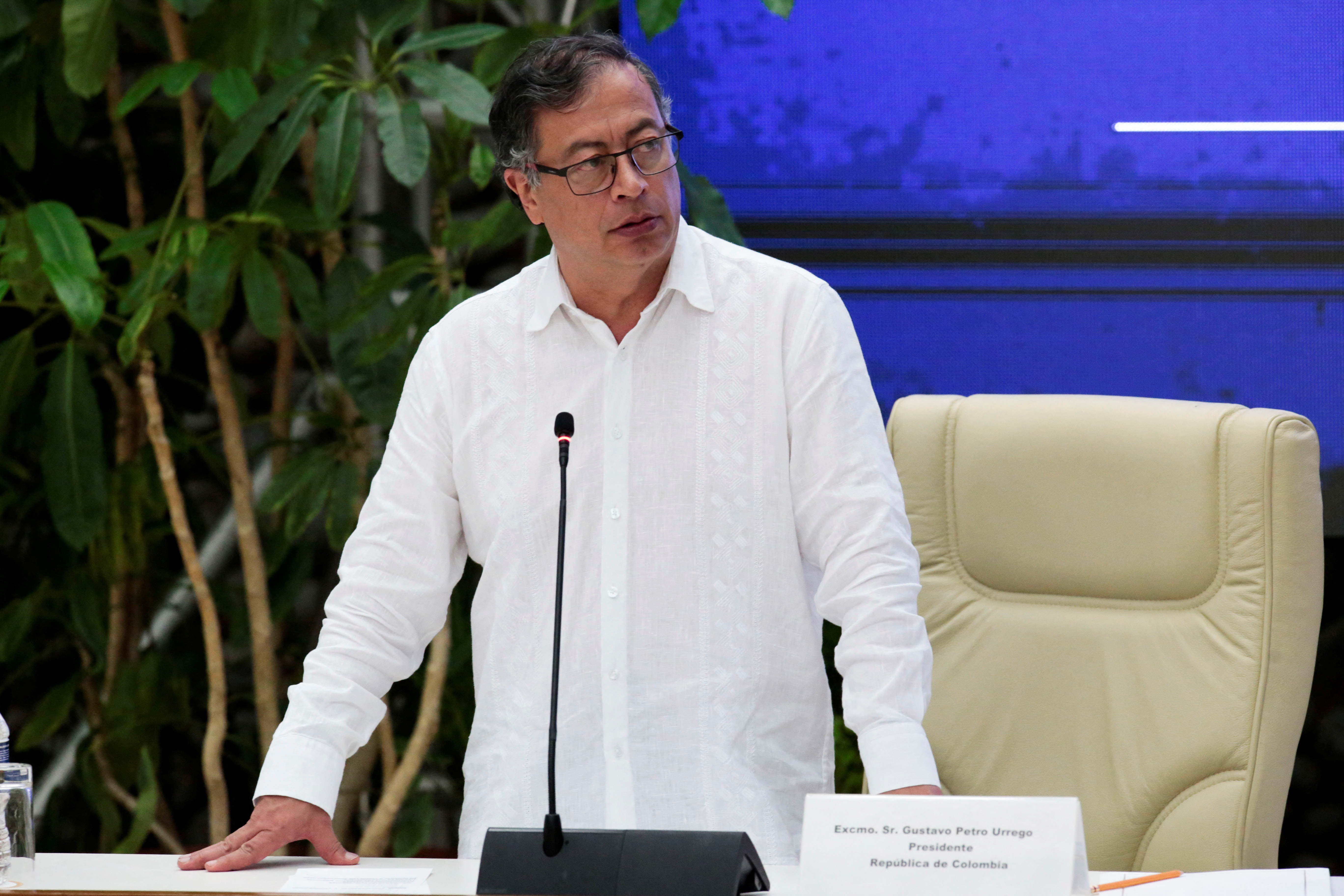 A man in a white shirt stands and speaks in front of a table set up with a microphone.