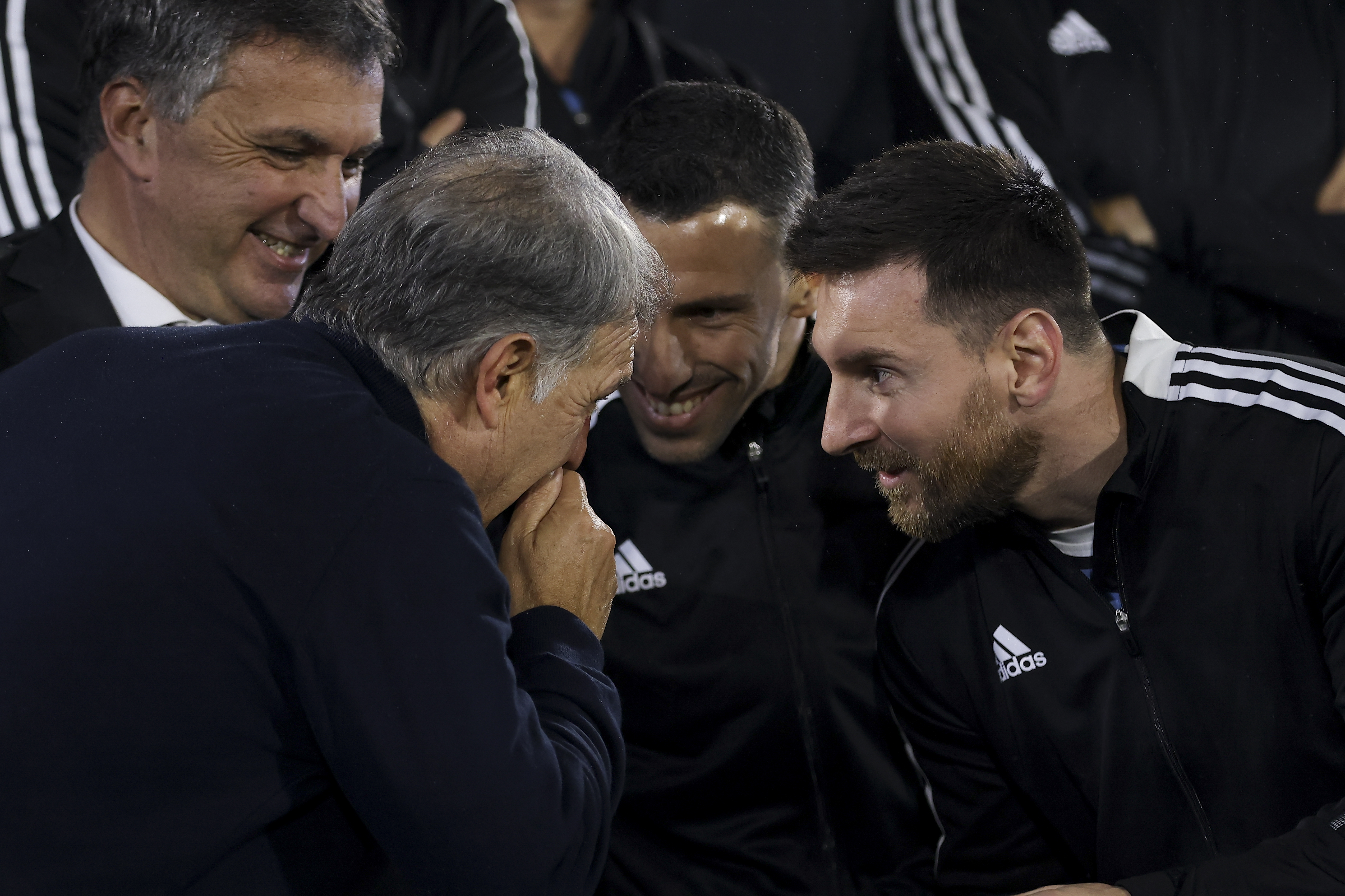 A man leans over, with hands cupped to his mouth, to speak with Lionel Messi, who also leans forward enthuisastically