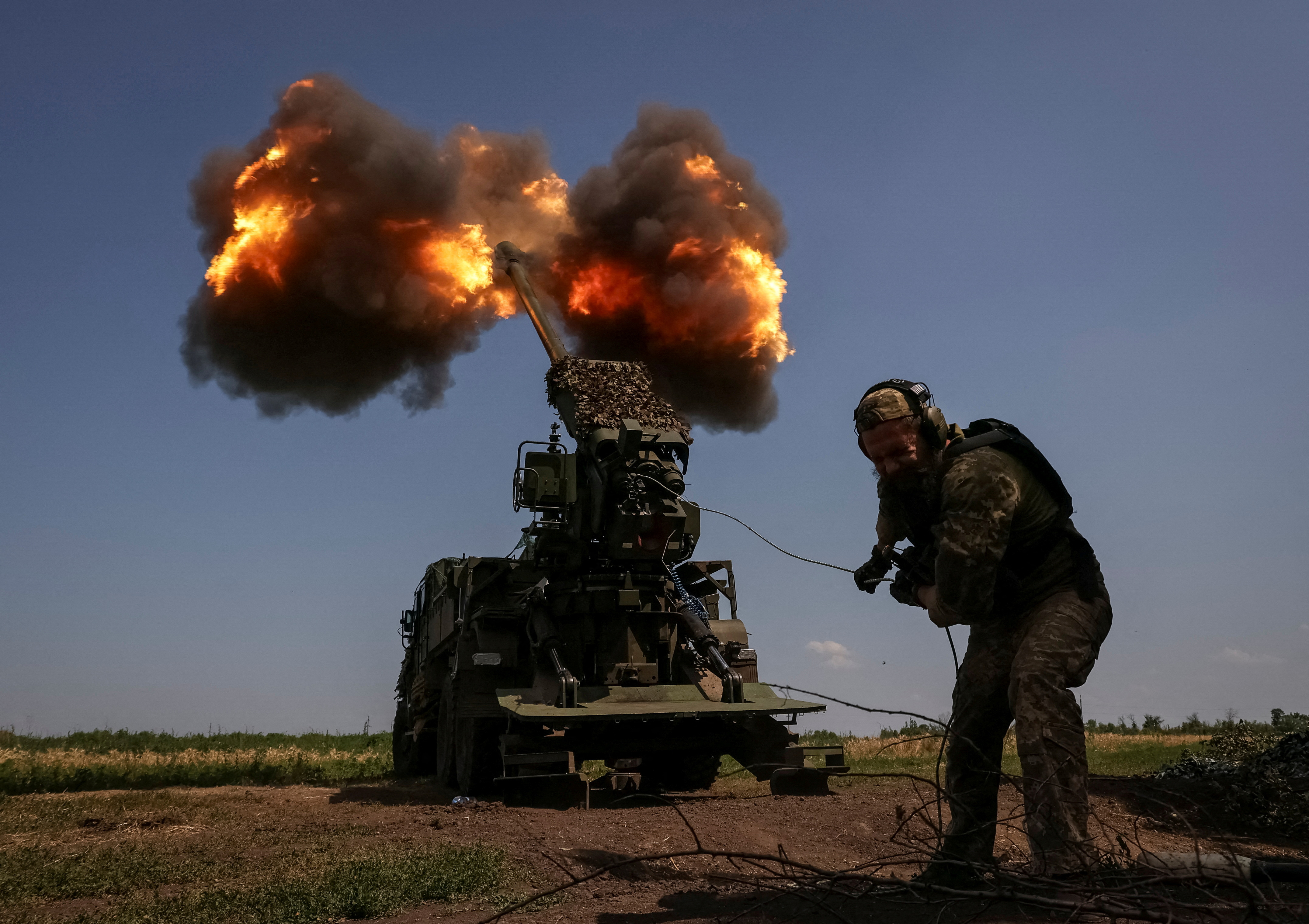 A Ukrainian soldier ducks as he fires a howitzer towards Russian lines near Bakhmut. The weapon is behind him and there are clouds of smoke and flames
