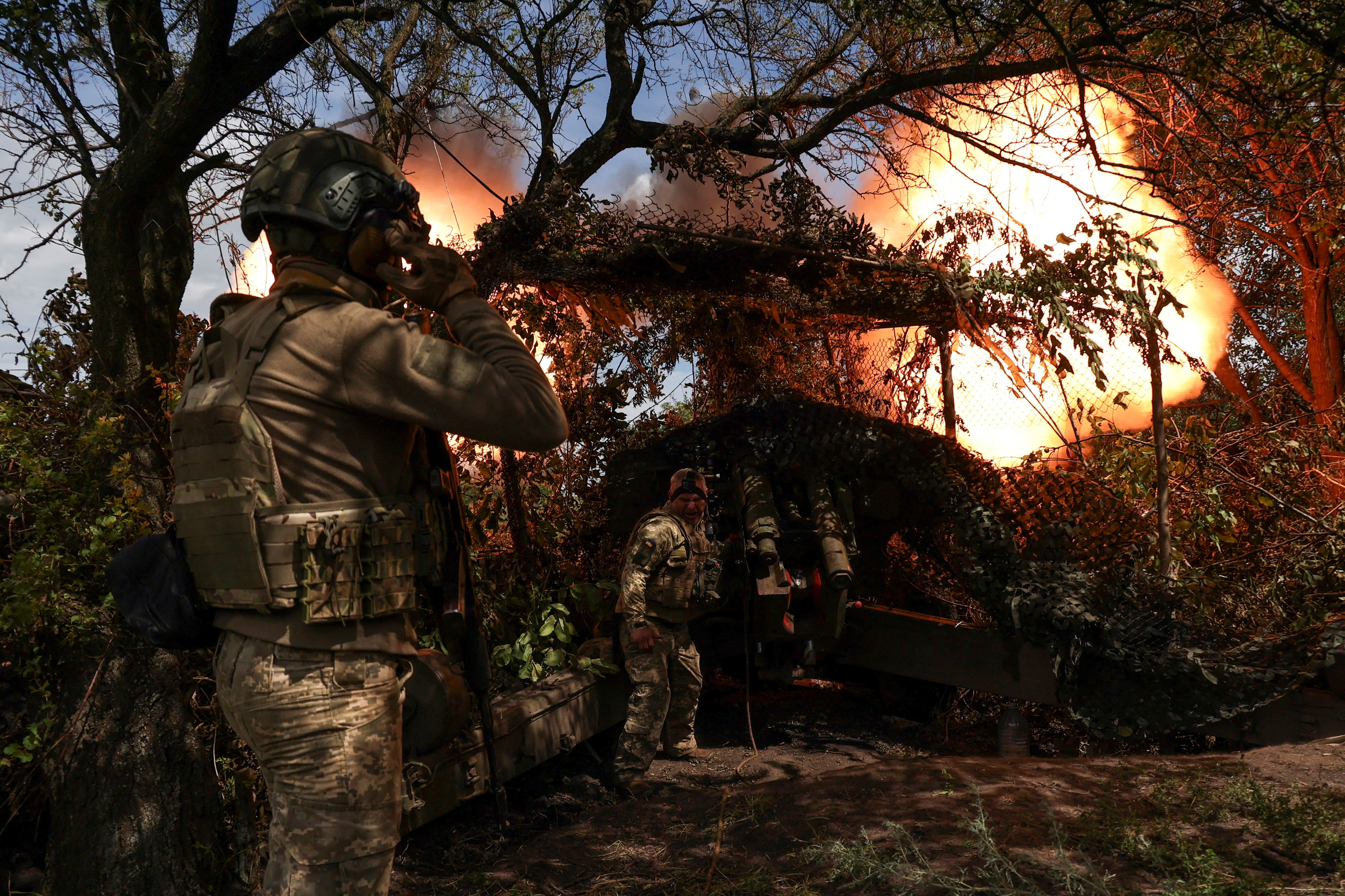 Ukrainian soldiers firing a howitzer. They are under the cover of trees. There's a large orange ball of flame from the weapon