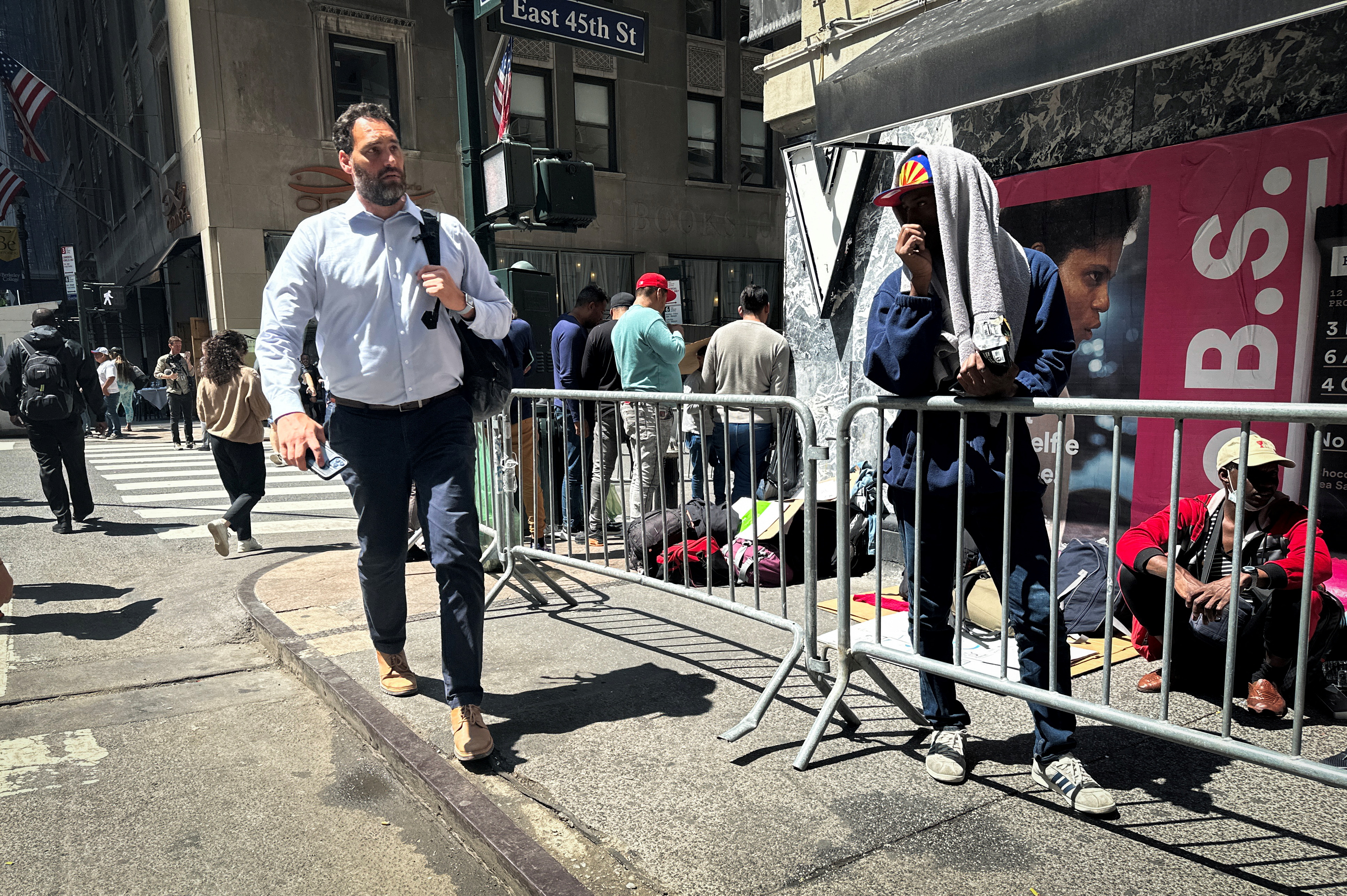 People walk past Recently arrived migrants to New York City waiting on the sidewalk outside the Roosevelt Hotel in midtown, Manhattan, where a temporary reception center has been established in New York City, New York, U.S., August 1, 2023. Some of the people in line sit on the sidewalk. Another stands at the metal barricade, separating the asylum seekers from the pedestrians.