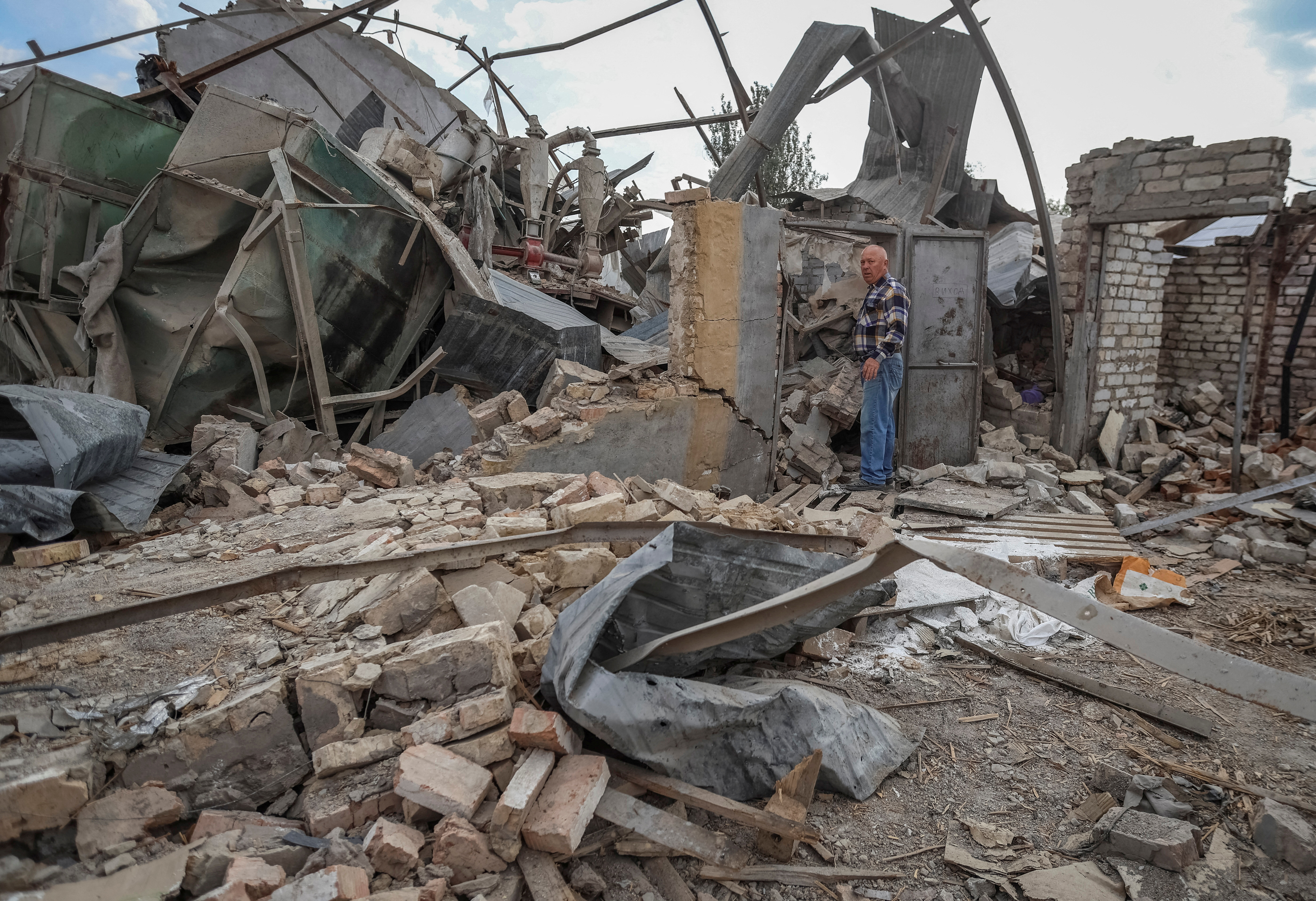 A Ukrainian farmer inspecting what's left of his grain storage after a Russian air attack. There is rubble all around and twisted metal.