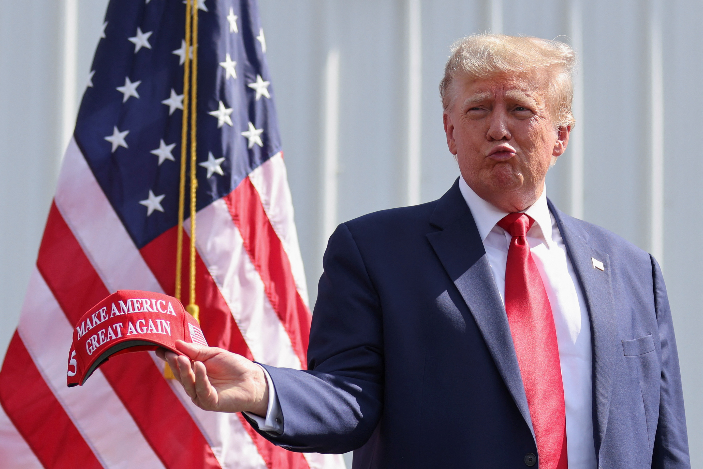 Former U.S. President and Republican presidential candidate Donald Trump attends a 2024 presidential election campaign event at Sportsman Boats in Summerville, South Carolina, U.S. September 25, 2023. In the photo, Trump — wearing a suit and red tie — pouts his mouth and holds a "Make America Great Again" baseball cap in front of an American flag.
