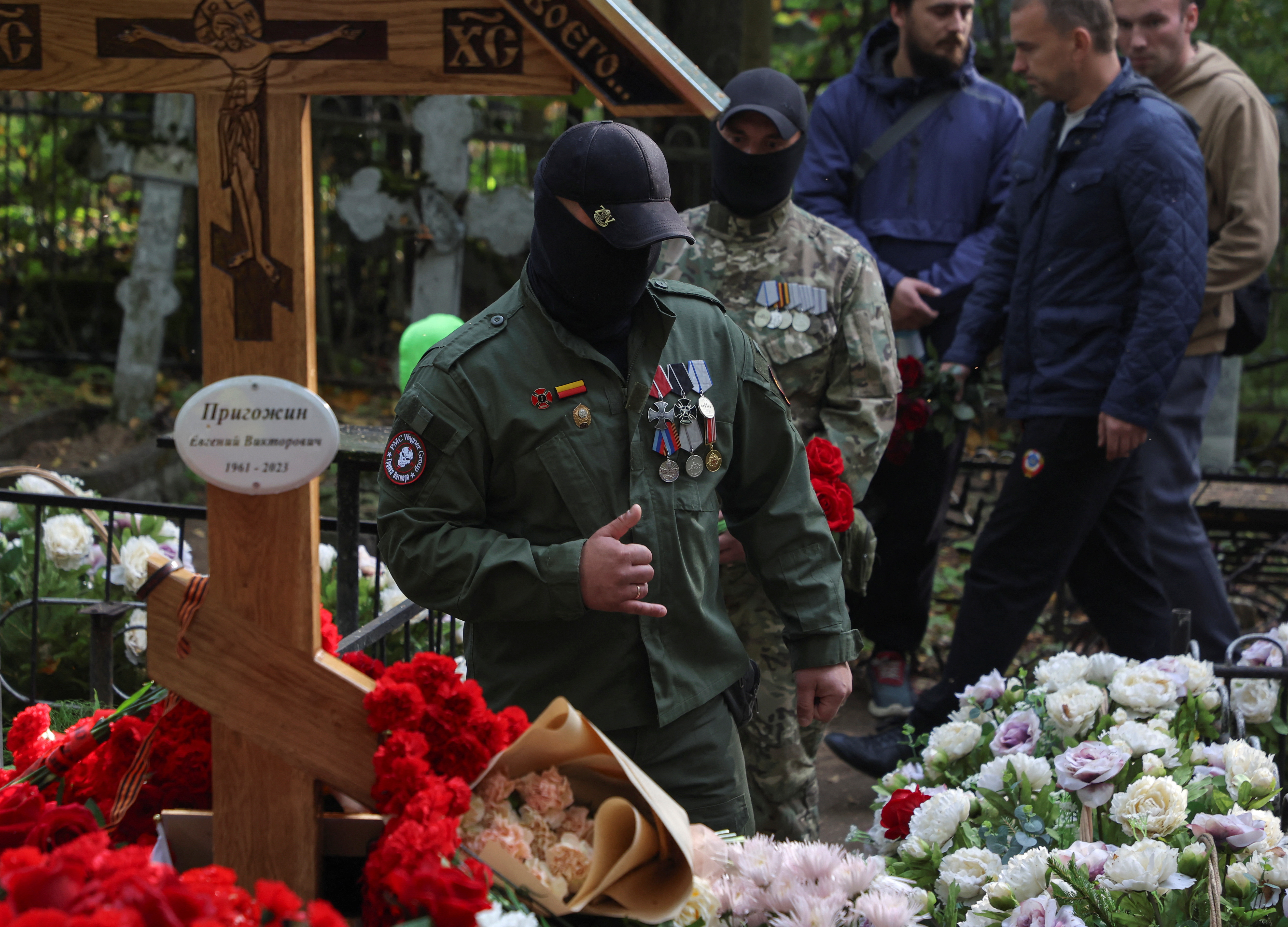 Wagner fighters at Prigozhin's grave. They are in fatigues and have covered their mouths with black masks. There are flowers around the grave.