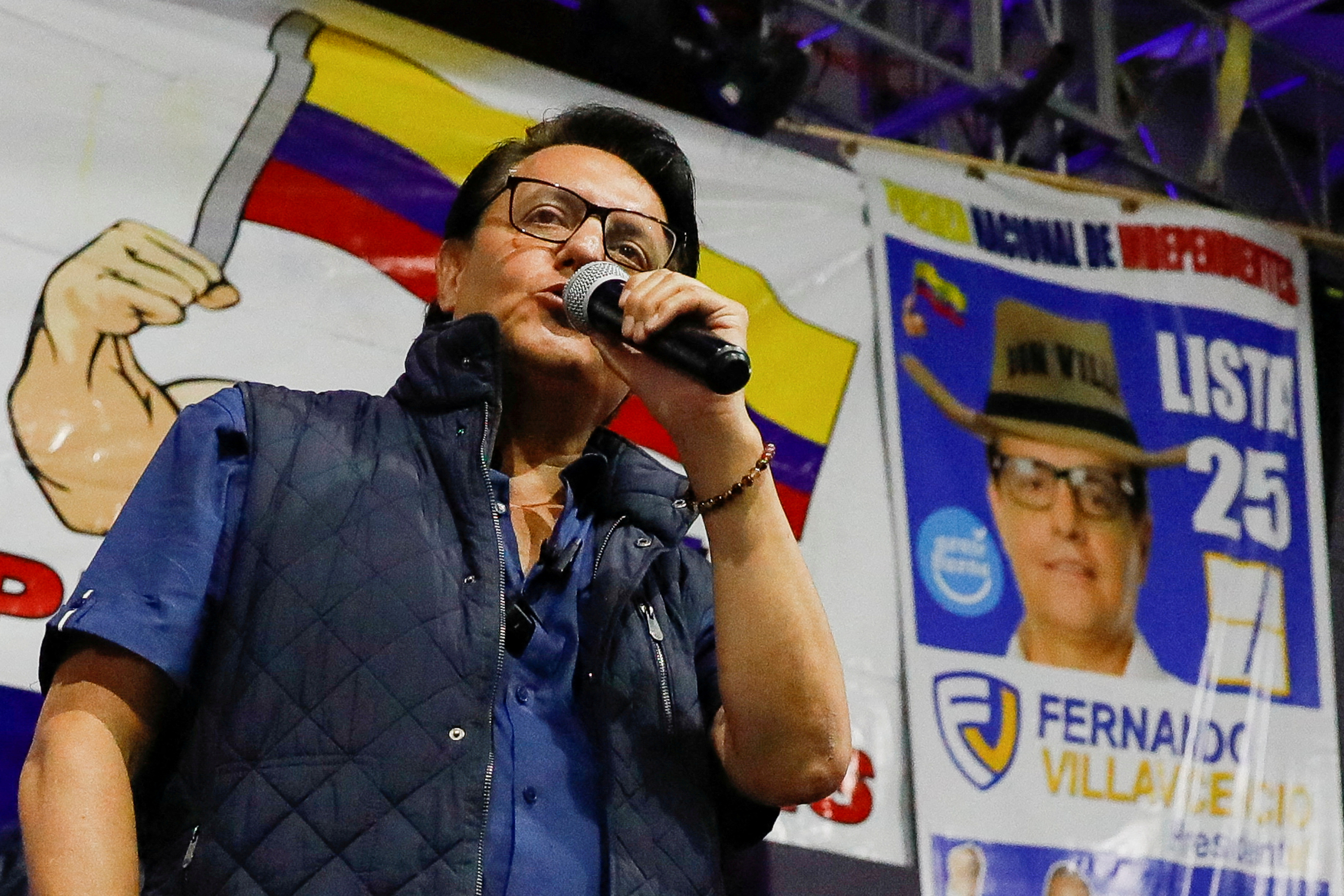 Fernando Villavincencio speaks into a microphone, wearing a navy blue puffer vest and blue shirt. Behind him is a banner featuring the Ecuadorian flag.