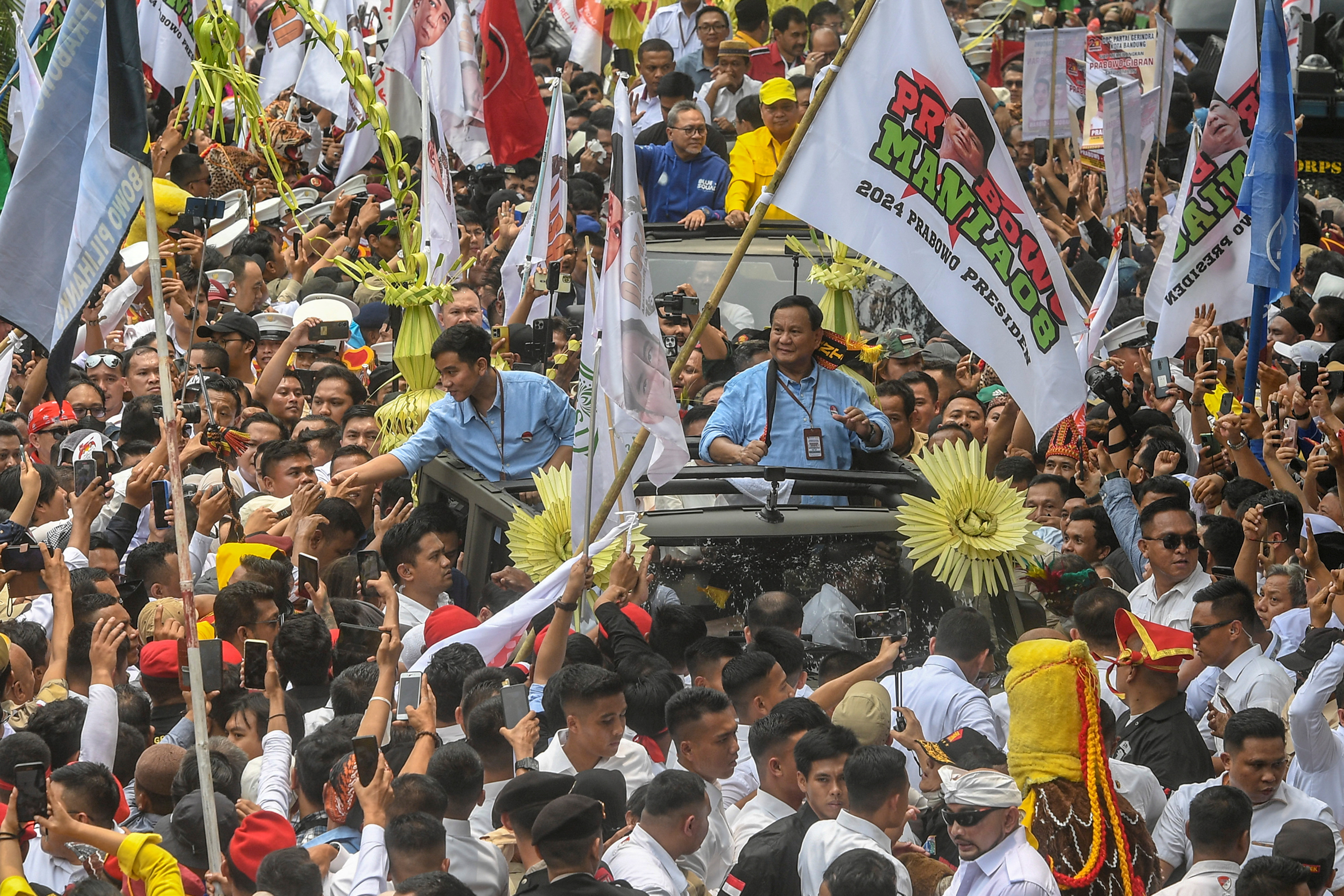 Supporters turn out to cheer Prabowo and Gibran as they travel through the streets of Jakarta. There are banners and flags. The two men are on an open top vehicle. GIbran is reaching out to shake hands with the crowds. 
