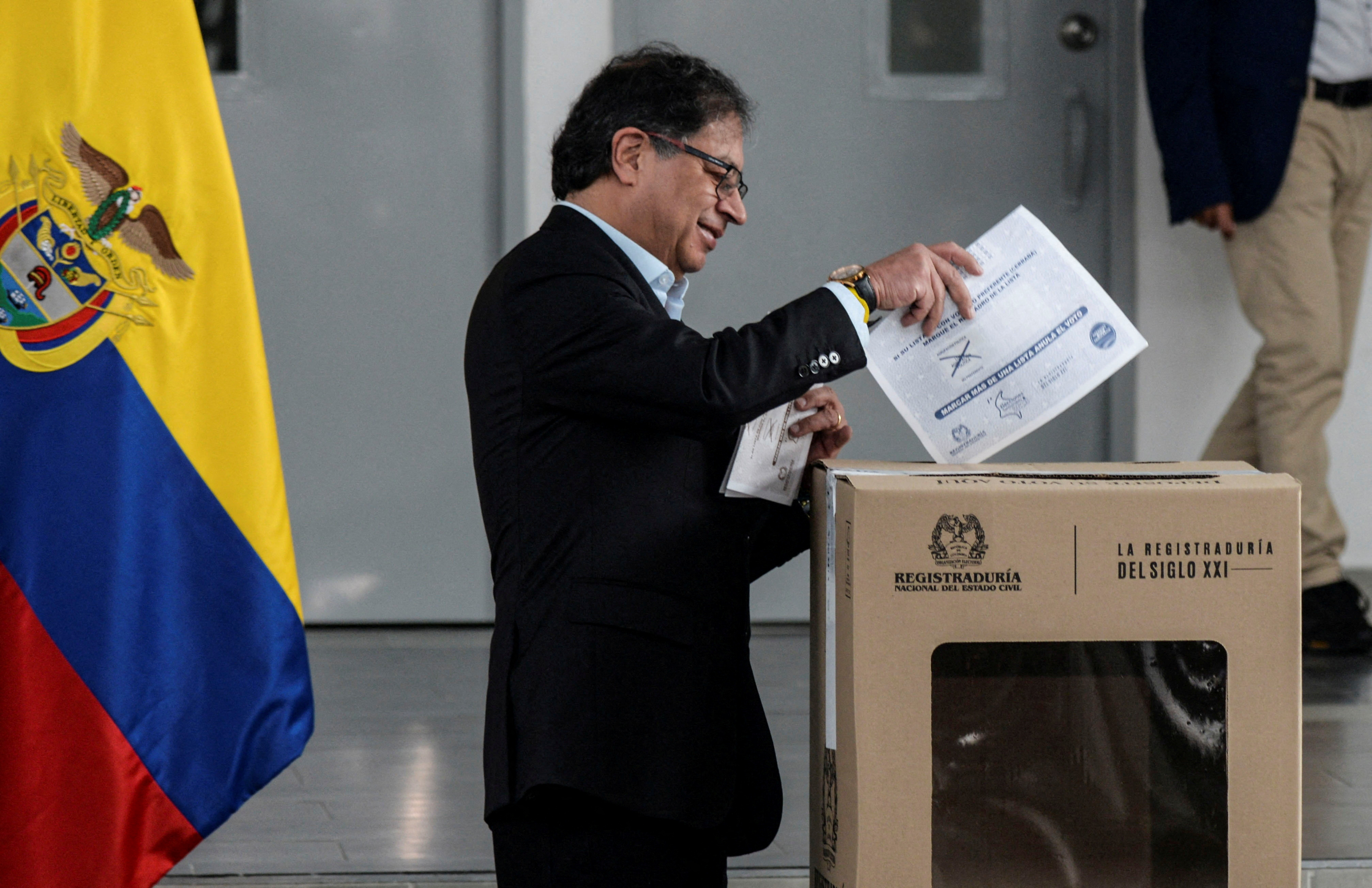 President Gustavo Petro, dressed in a dark suit, puts a ballot into a cardboard box in a nondescript room. The Colombian flag is behind him.