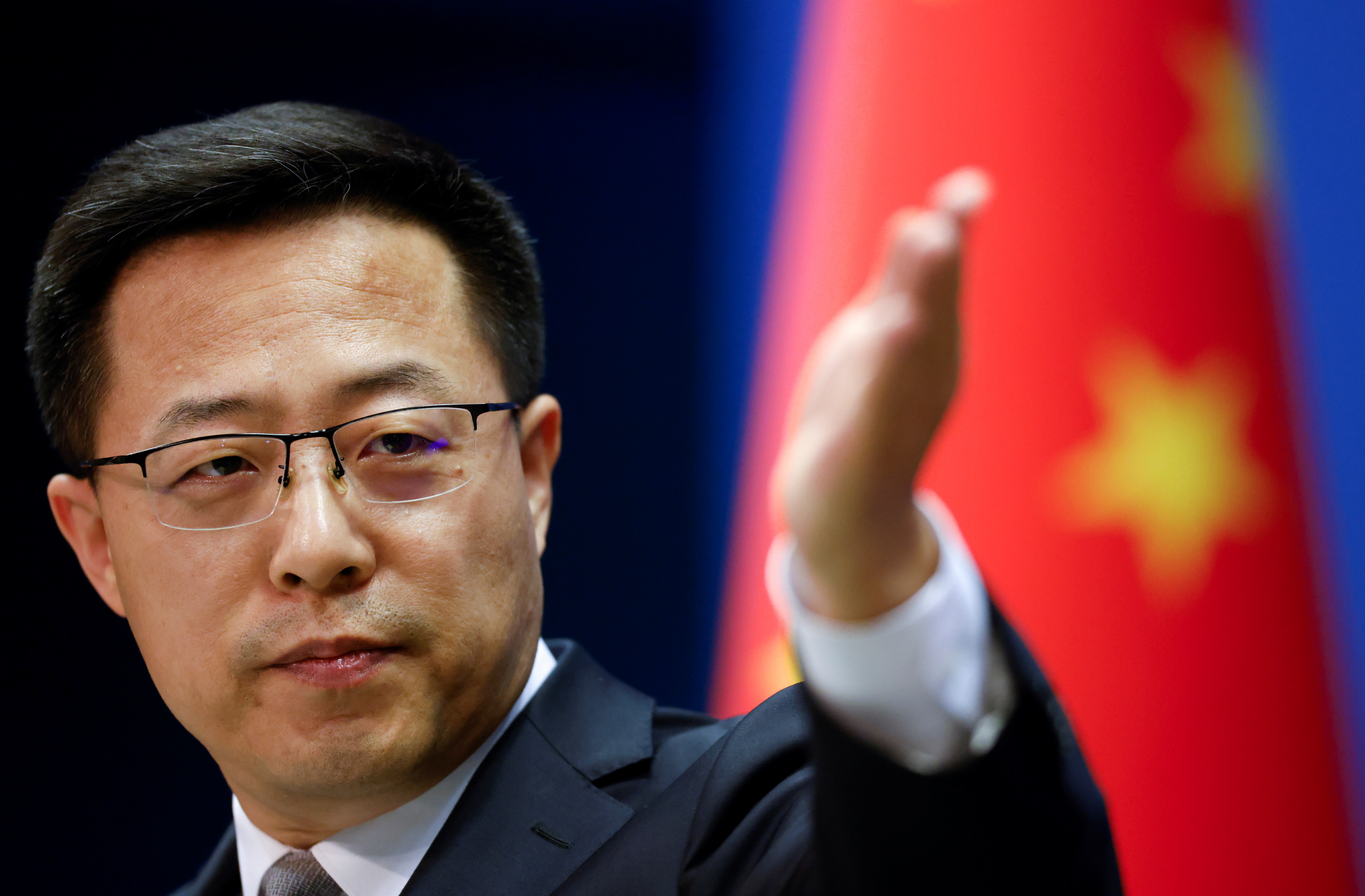 Zhao Lijian taking a question at a press conference. A Chinese flag is behind him. He has his hand out 