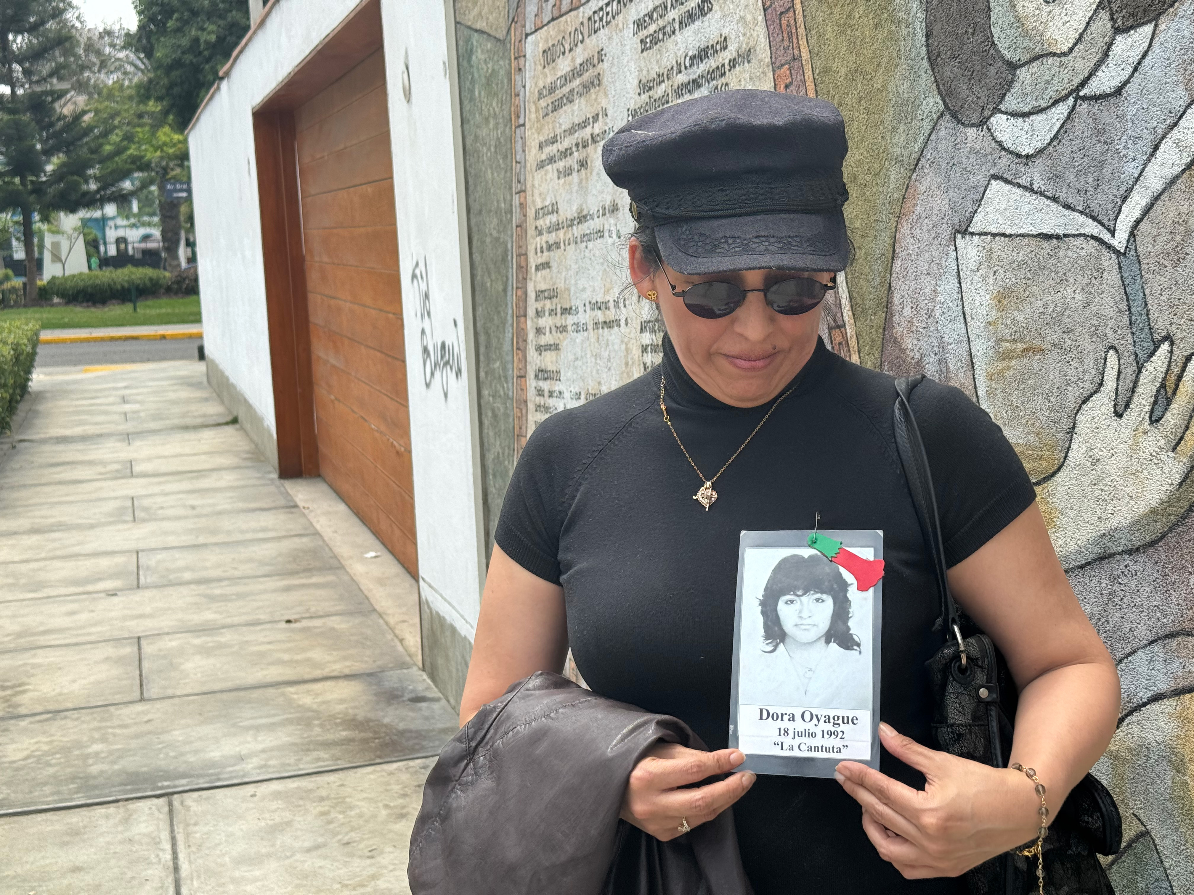 A woman in a black cap and black shift looks down at the black-and-white photo of her late sister that she cradles in her hands.