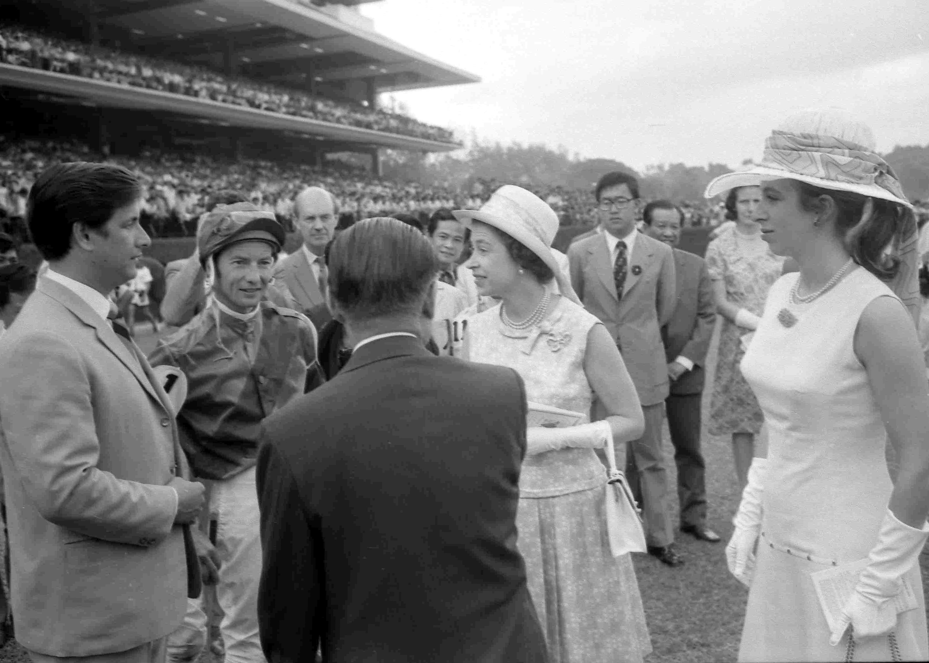 Queen Elizabeth Ii at the Singapore Turf Club in 1972. She is speaking to jockey Lester Piggott. Her daughter Princess Anne is with her. The grandstand is behind them.