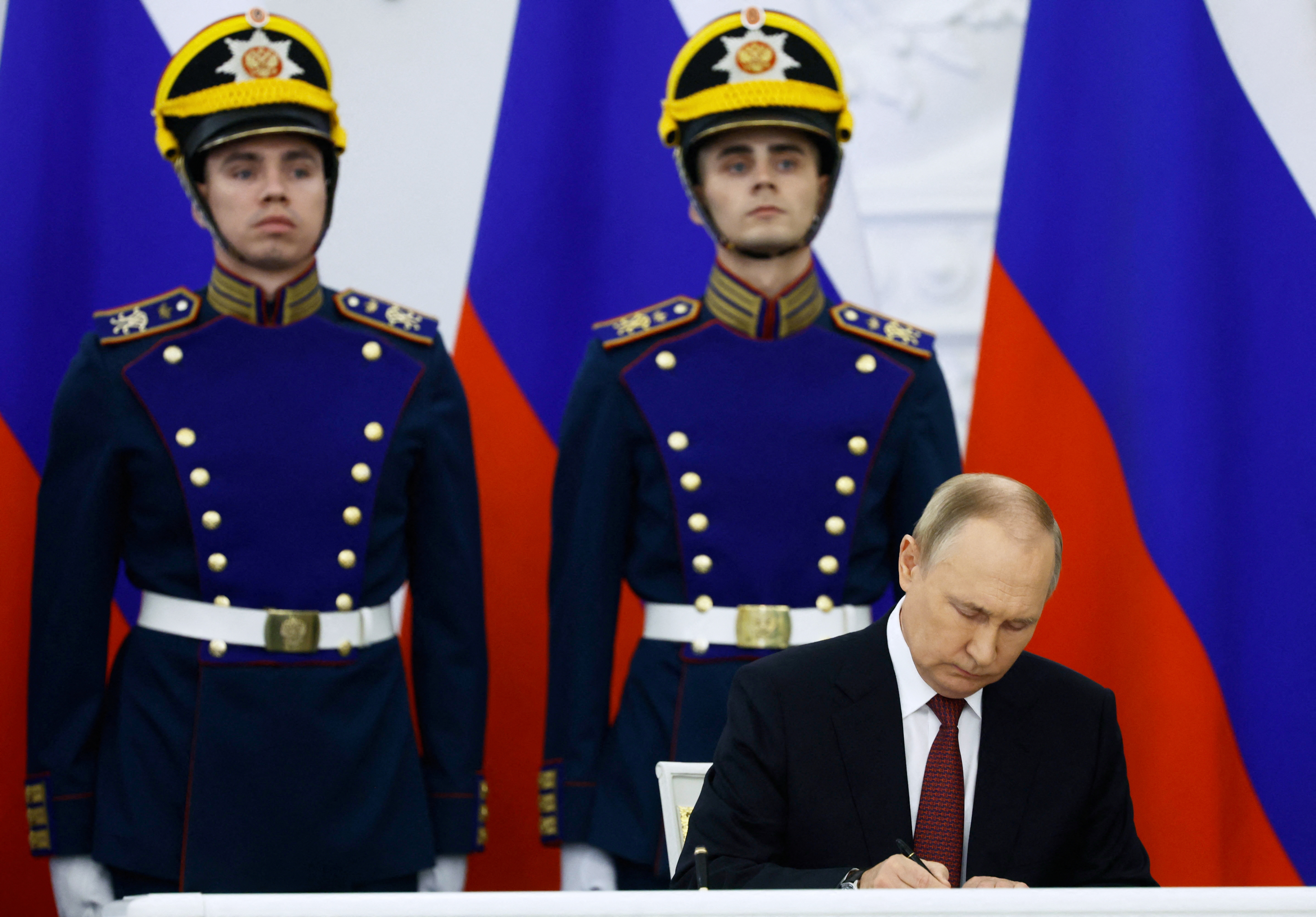 Two soldiers in ceremonial uniform standing to attention with Russian flags behind them. Putin is in front signing the 'annexation' of four Ukrainian territories partly occupied by Russia.