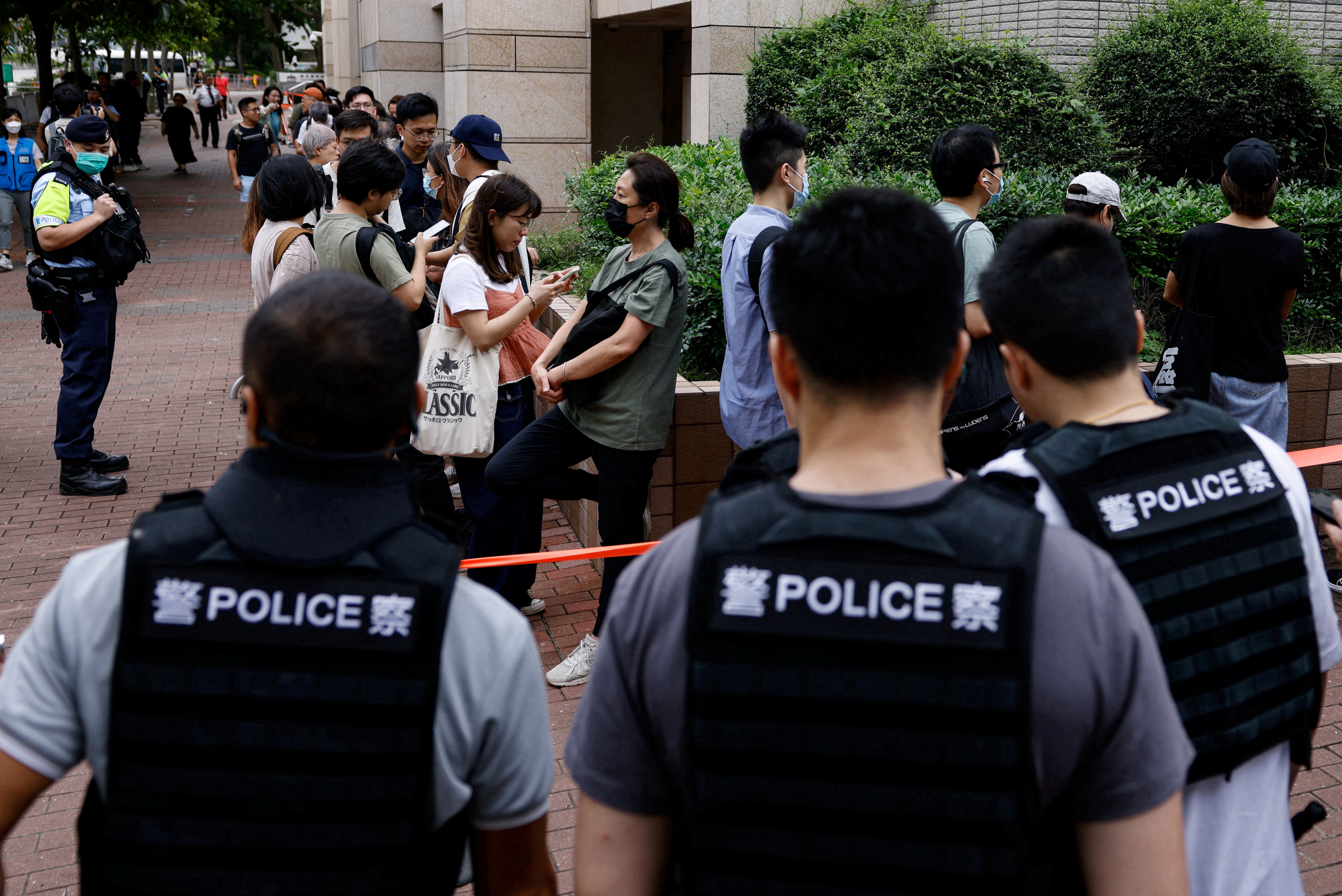 Police officers keep watch outside the court. There is a long queue of people waiting to go inside to hear the verdict.. The officers are wearing blue vests reading POLICE across their backs.