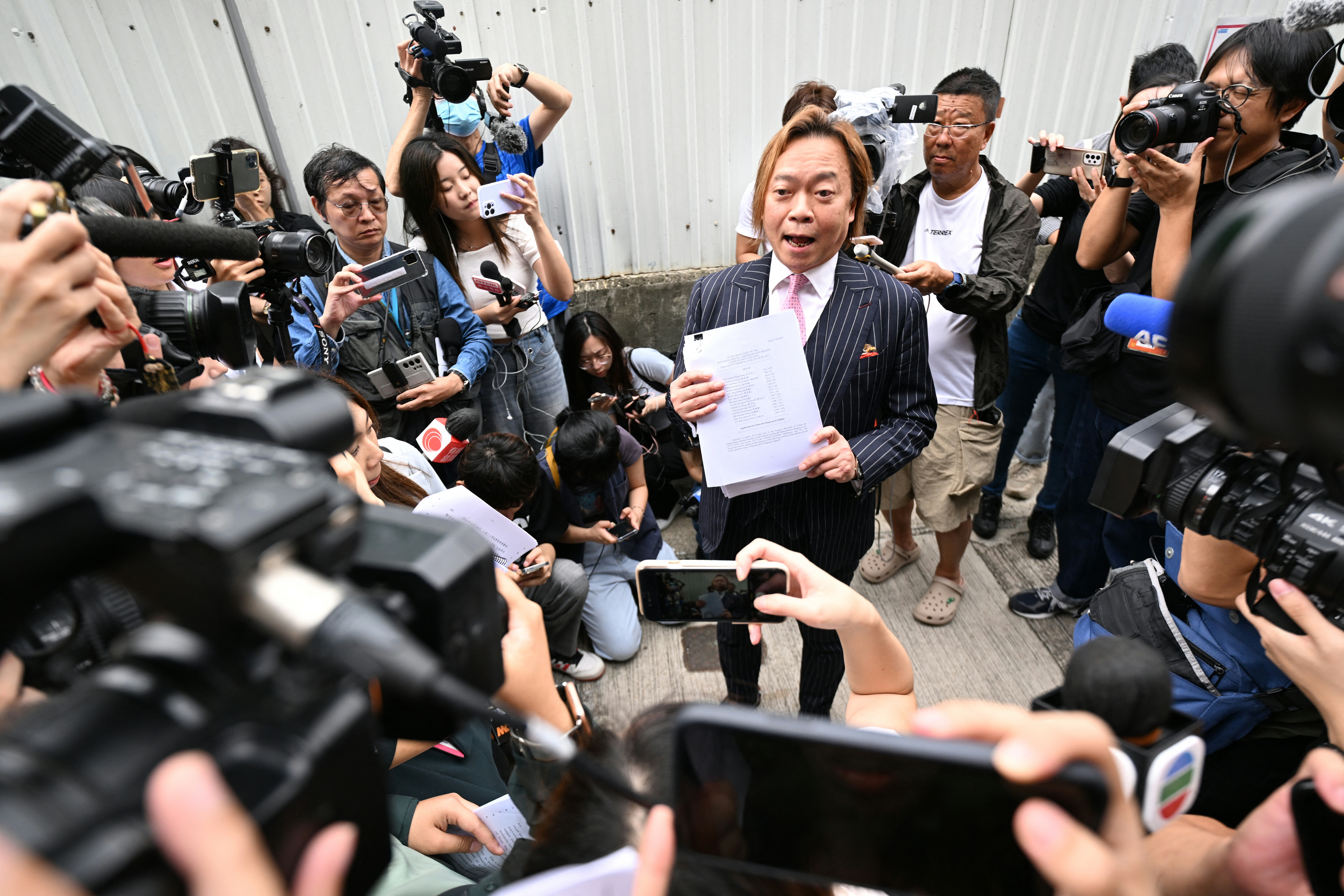 Lawrence Lau speaking to the media. He is wearing a pin-striped suit and holding a document. Reporters are all around him. Some with cameras.