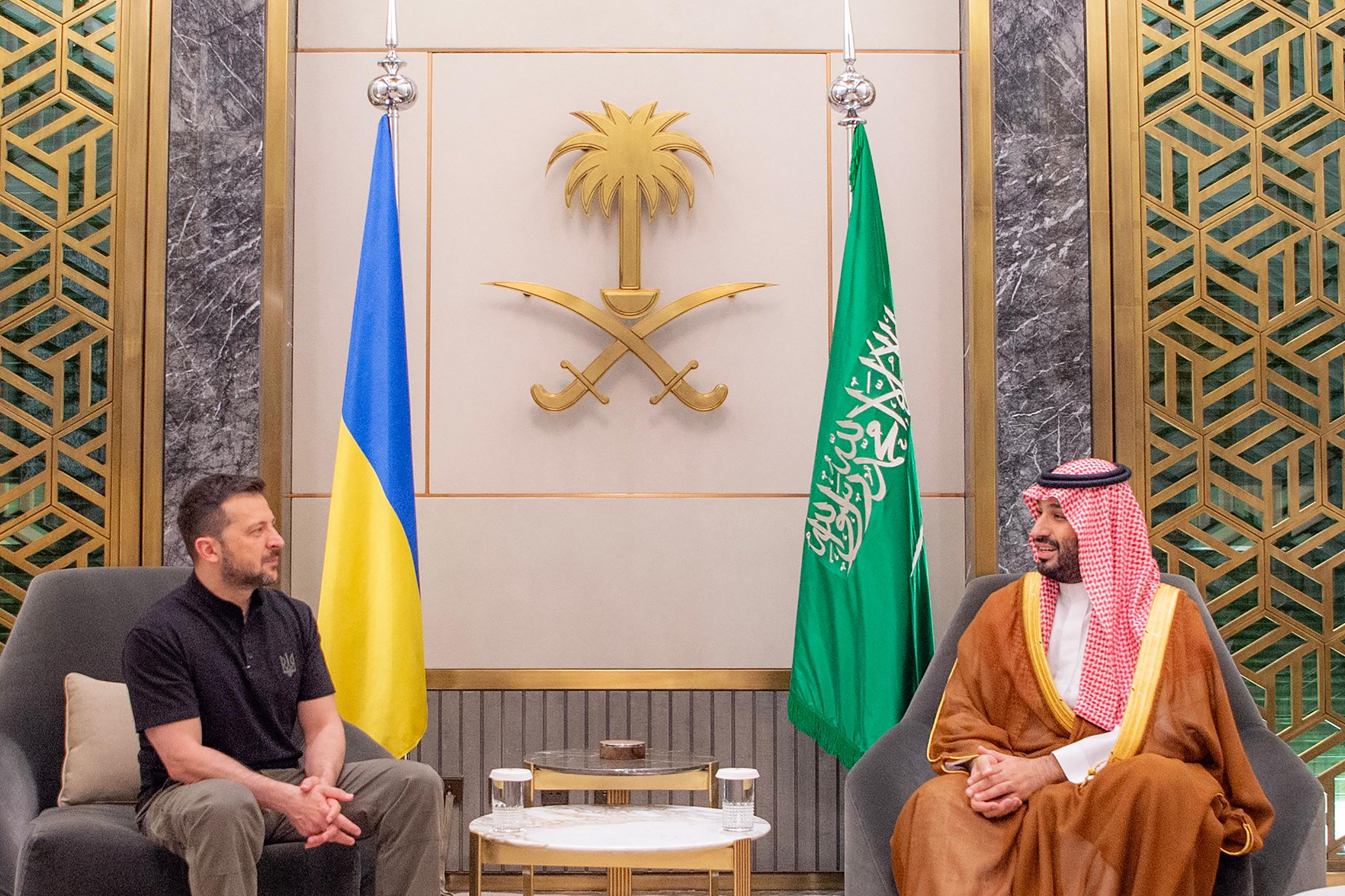 Volodymyr Zelenskyy at a meeting with Saudi Crown Prince Mohammed Bin Salman. They are seated either side of a coffee table in front of flags of their respective countries. Zelenskyy is in a black polo shirt and khaki trousers. Mohammed Bin Salman is in a traditional robe.