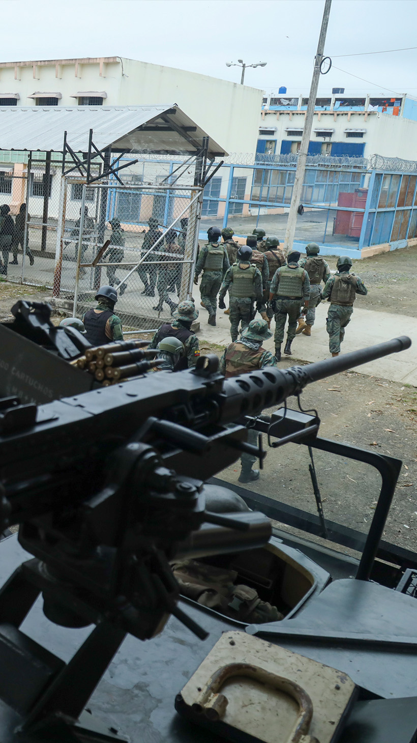 A soldier sits behind the sight of a rifle, looking out on the El Rodeo prison, its long white buildings and chain-link fence in the distance. A row of soldiers enters the prison below.
